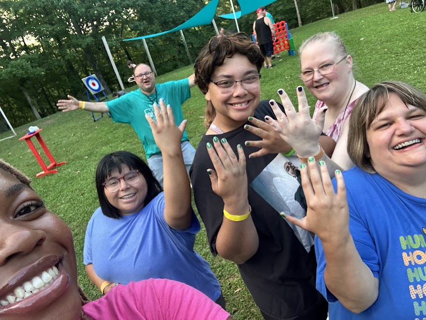 Group of smiling people outdoors, showing off painted nails, with a playground and trees in the background.