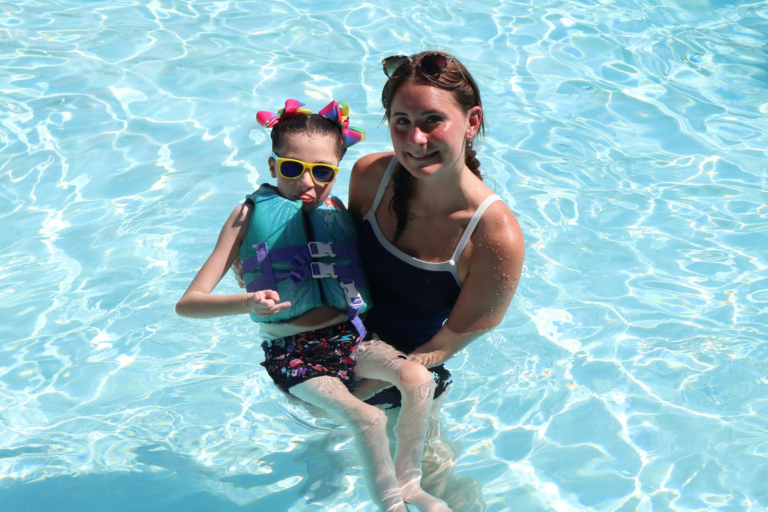 Woman and girl in a swimming pool, woman holding girl, both smiling, girl wearing sunglasses and colorful hair accessories.
