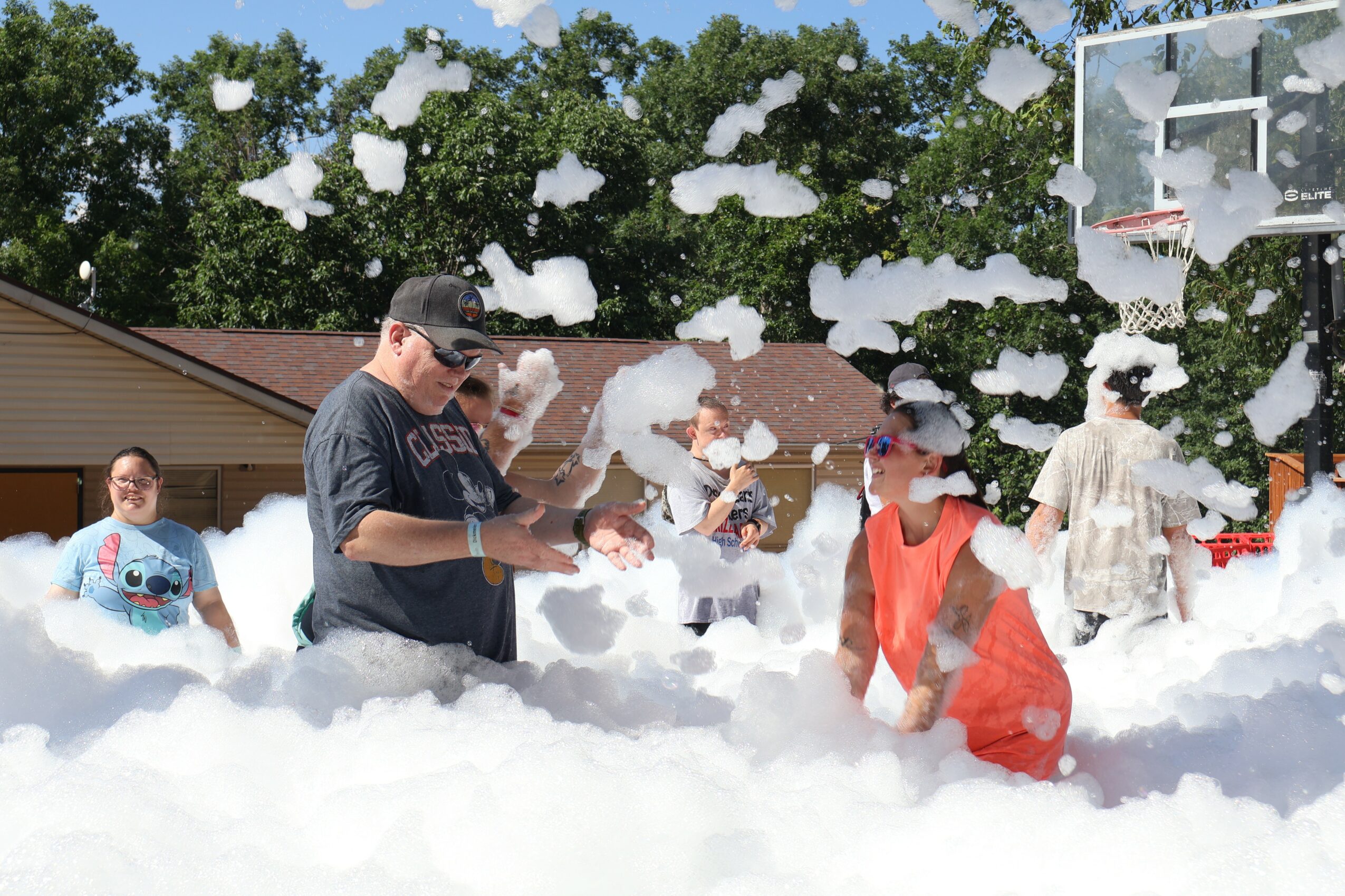 Group of children and an adult playing in foam outdoors near a house and basketball hoop.
