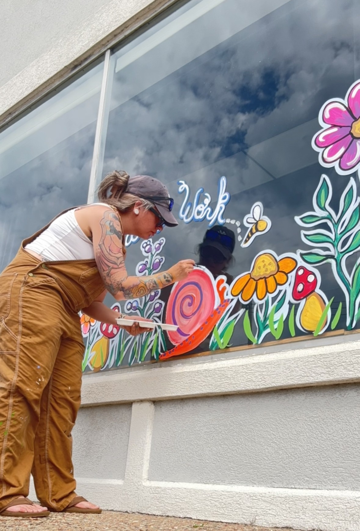 Woman painting colorful flowers and a snail on a window with clouds reflected in the glass.