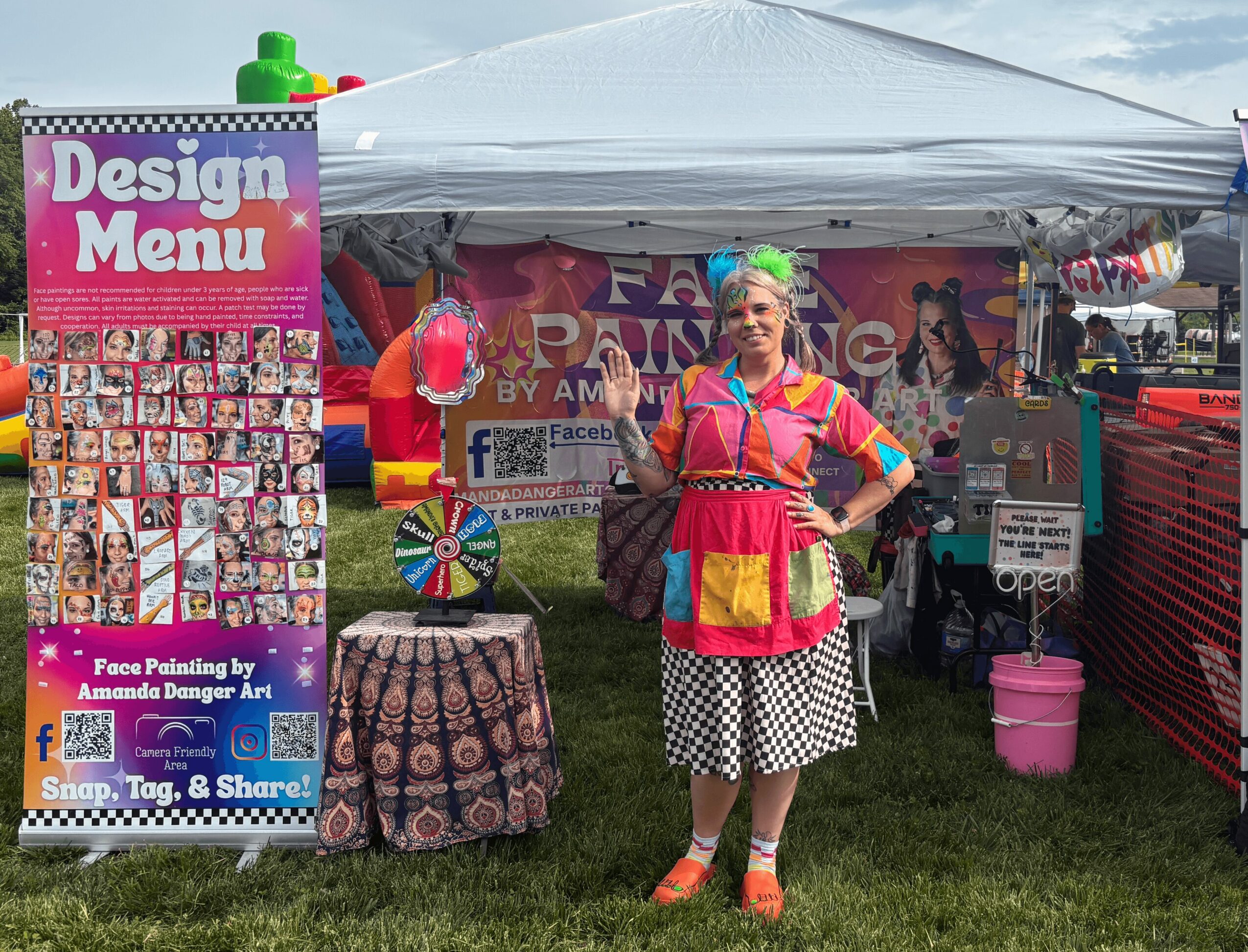 Woman in colorful outfit standing in front of a tent at an outdoor event, with a face painting sign and display board nearby.