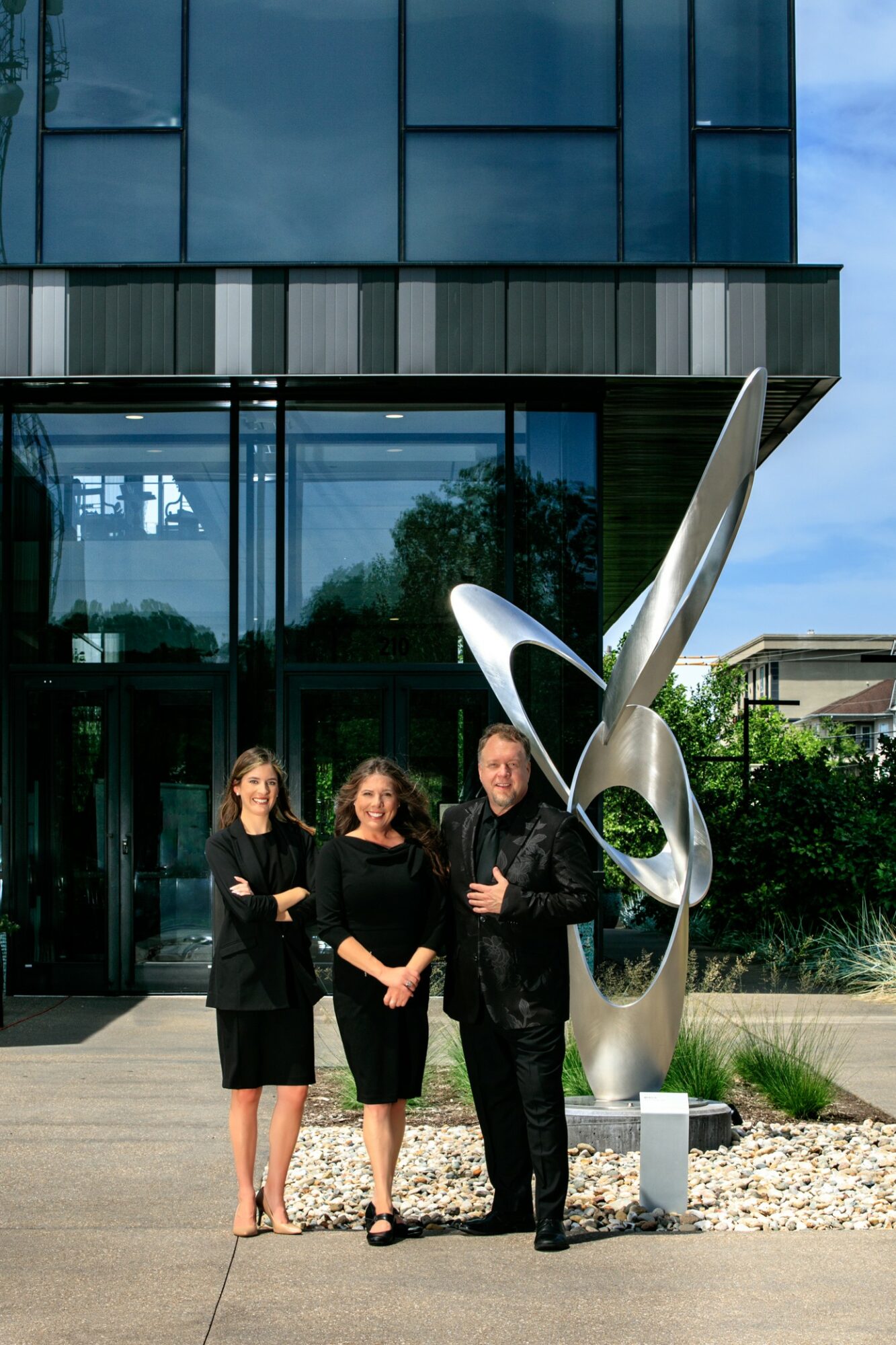 Three people stand in front of a modern building with glass windows and a large abstract sculpture, outdoors during daytime.