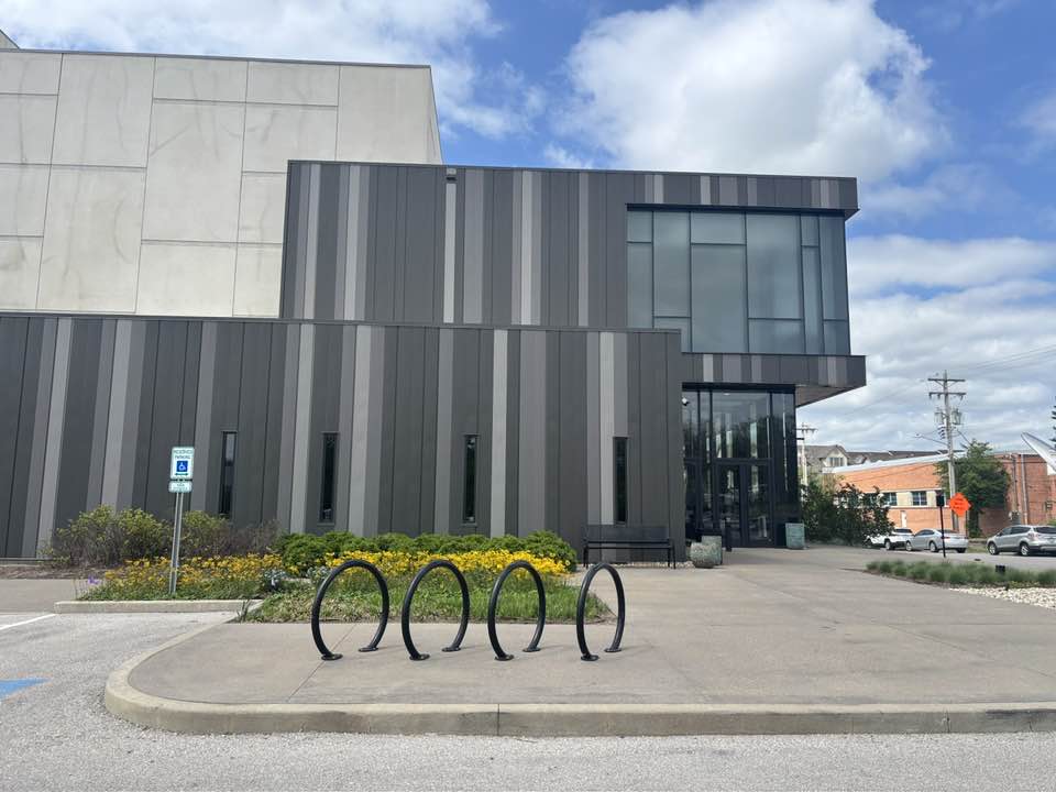 Modern building with dark vertical panels, glass entrance, and a parking lot with bike racks, under a partly cloudy sky.