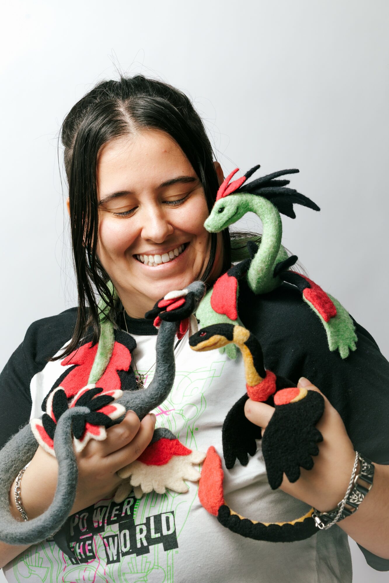 Smiling woman holding colorful dragon plush toys, wearing a black jacket and a white shirt, against a plain background.
