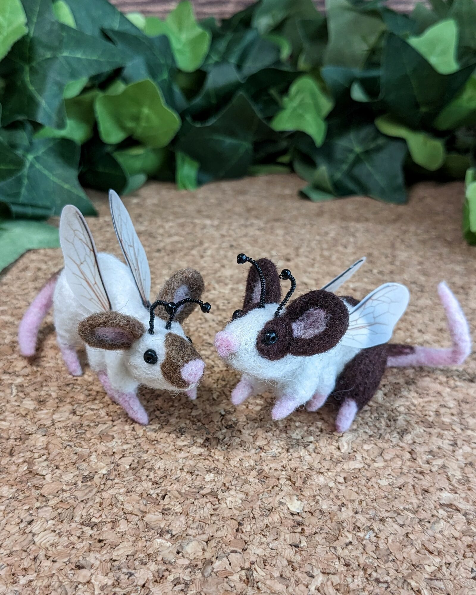 Two felt mice with wings and antennae on a cork surface, surrounded by green leaves, facing each other.