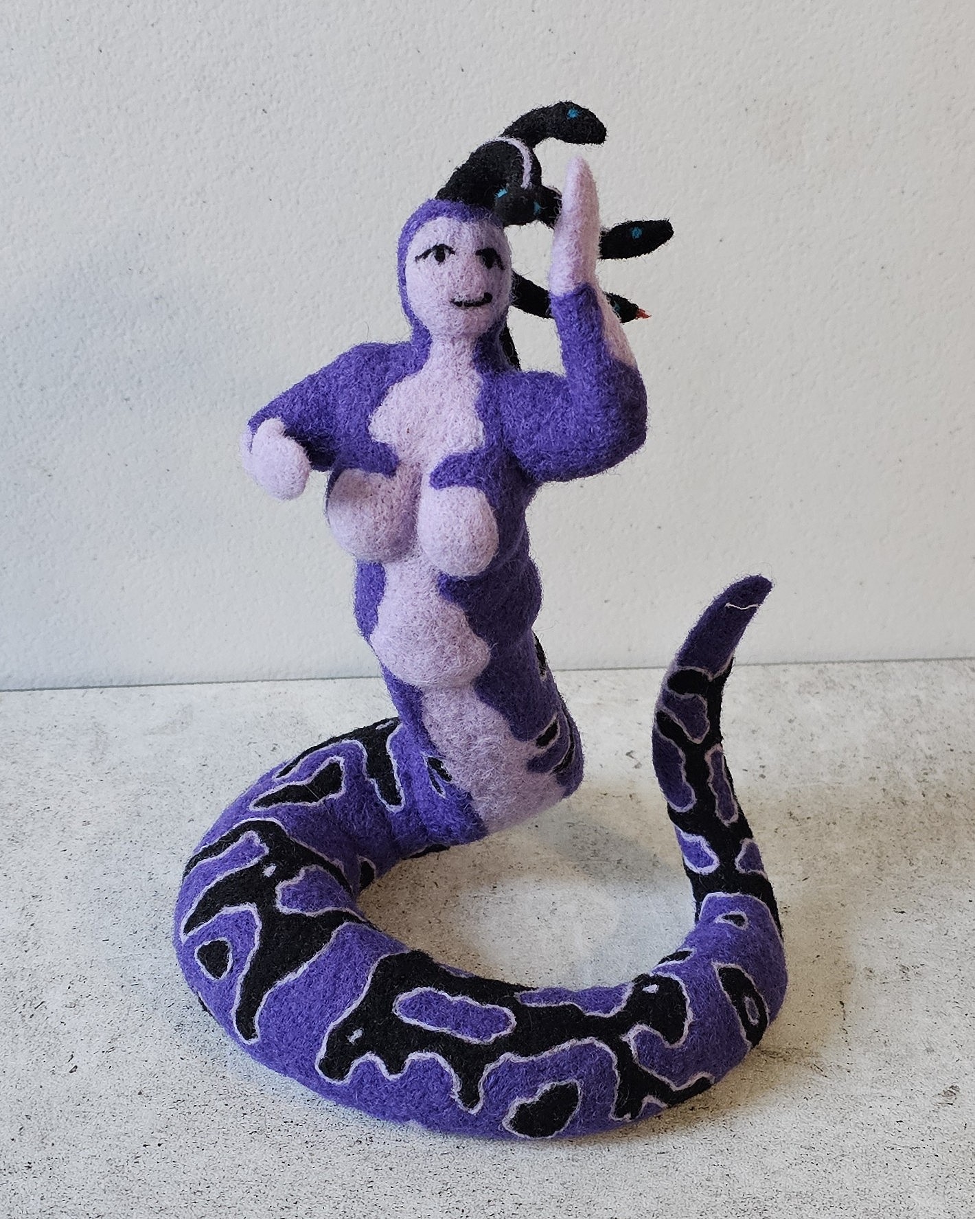 Purple and white plush snake with a smiling face and a hat, standing upright against a plain wall.