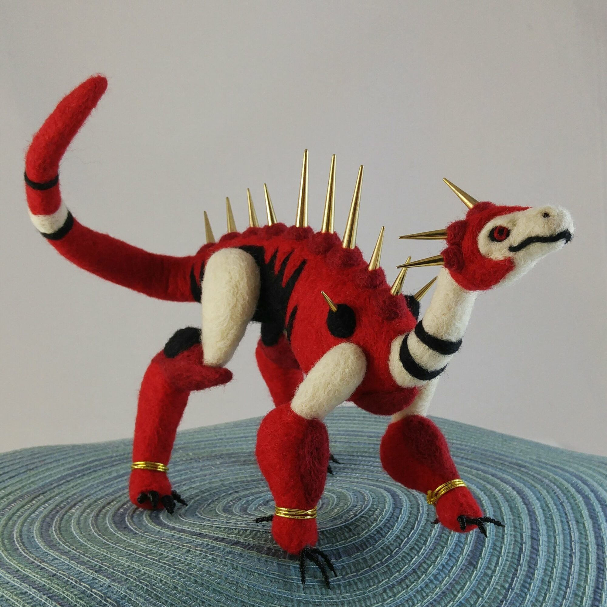 Red plush creature with spikes, striped limbs, and a long tail, standing on a textured surface against a plain background.