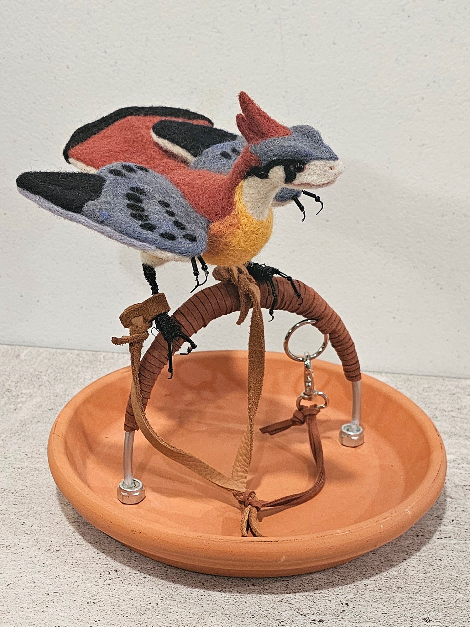 Colorful bird-shaped plush toy perched on a stand inside a round wooden dish, with a white background.