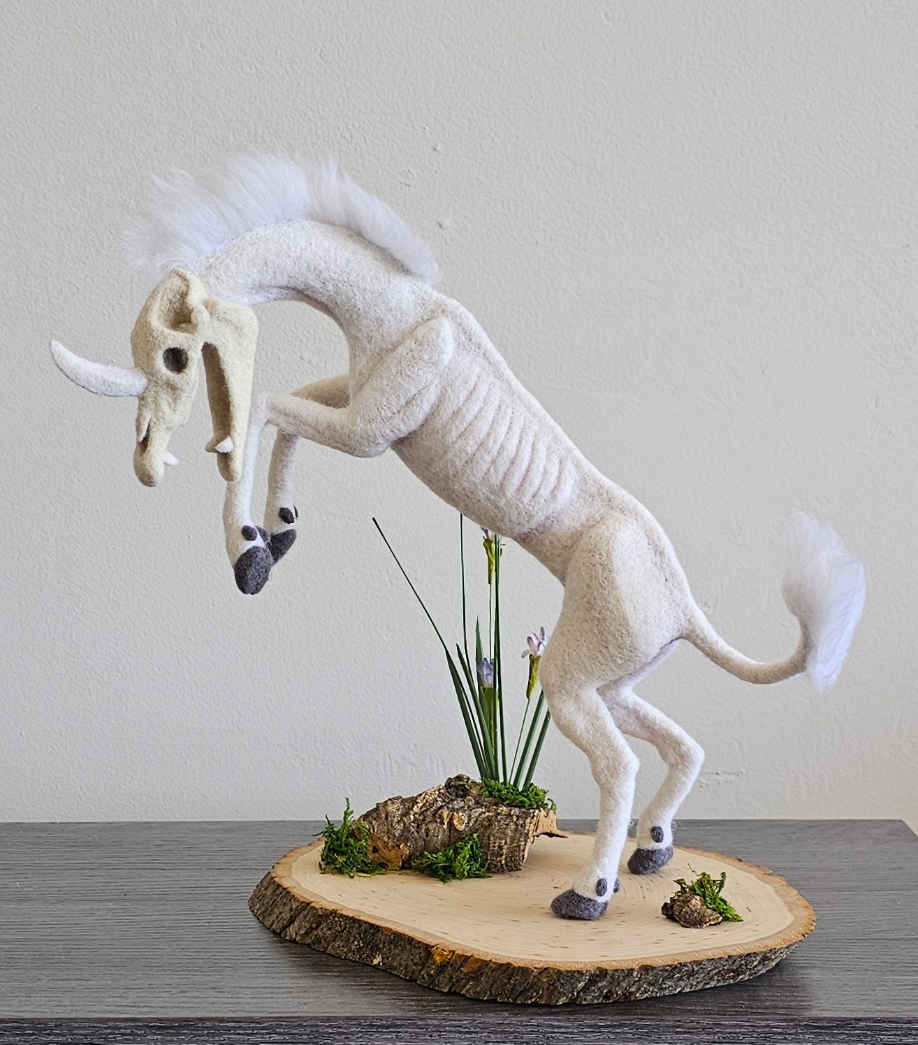 White plush horse with a flowing mane and tail, standing on a wooden base with plants and rocks, against a plain background.
