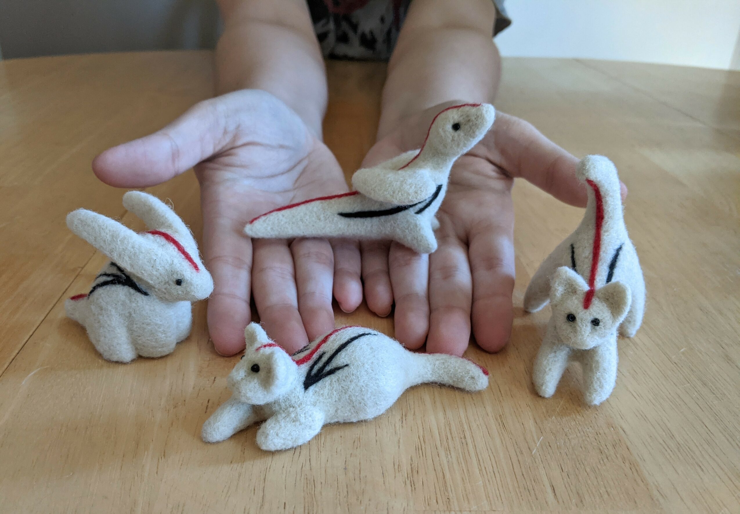 Two hands hold plush animal toys resembling rabbits and a snake, with four plush animals on a wooden surface.