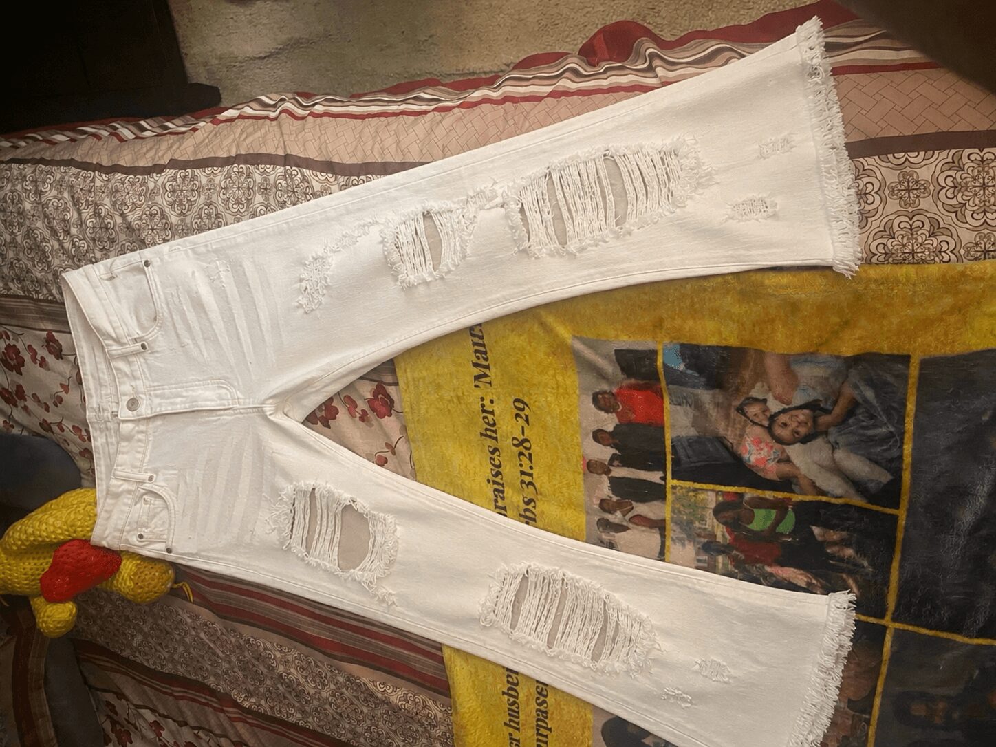 White distressed jeans with multiple rips, laid on a bed with a colorful blanket and a magazine underneath.