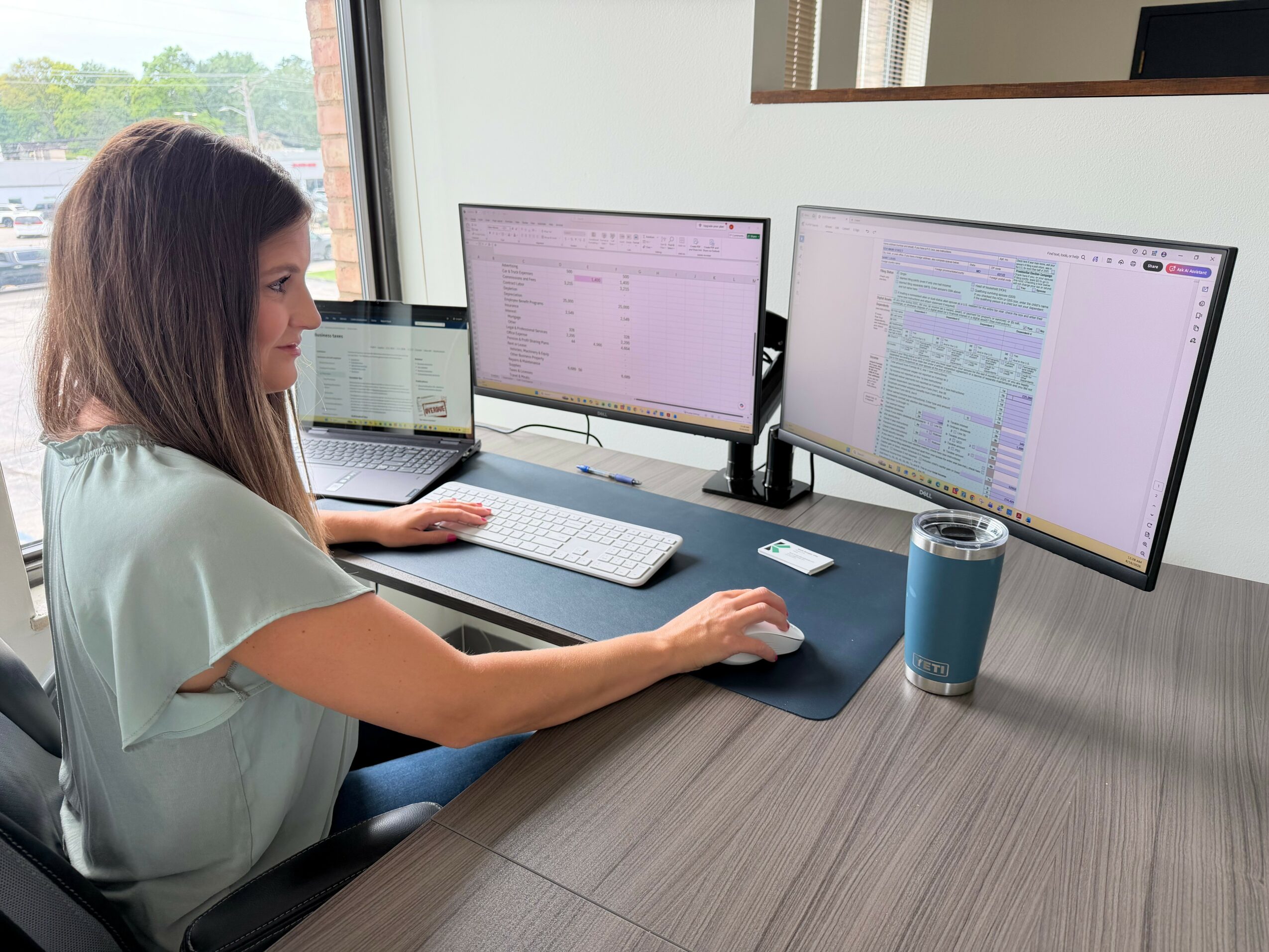 Woman working at a desk with three monitors, a laptop, and a tumbler, near a window with a view outside.