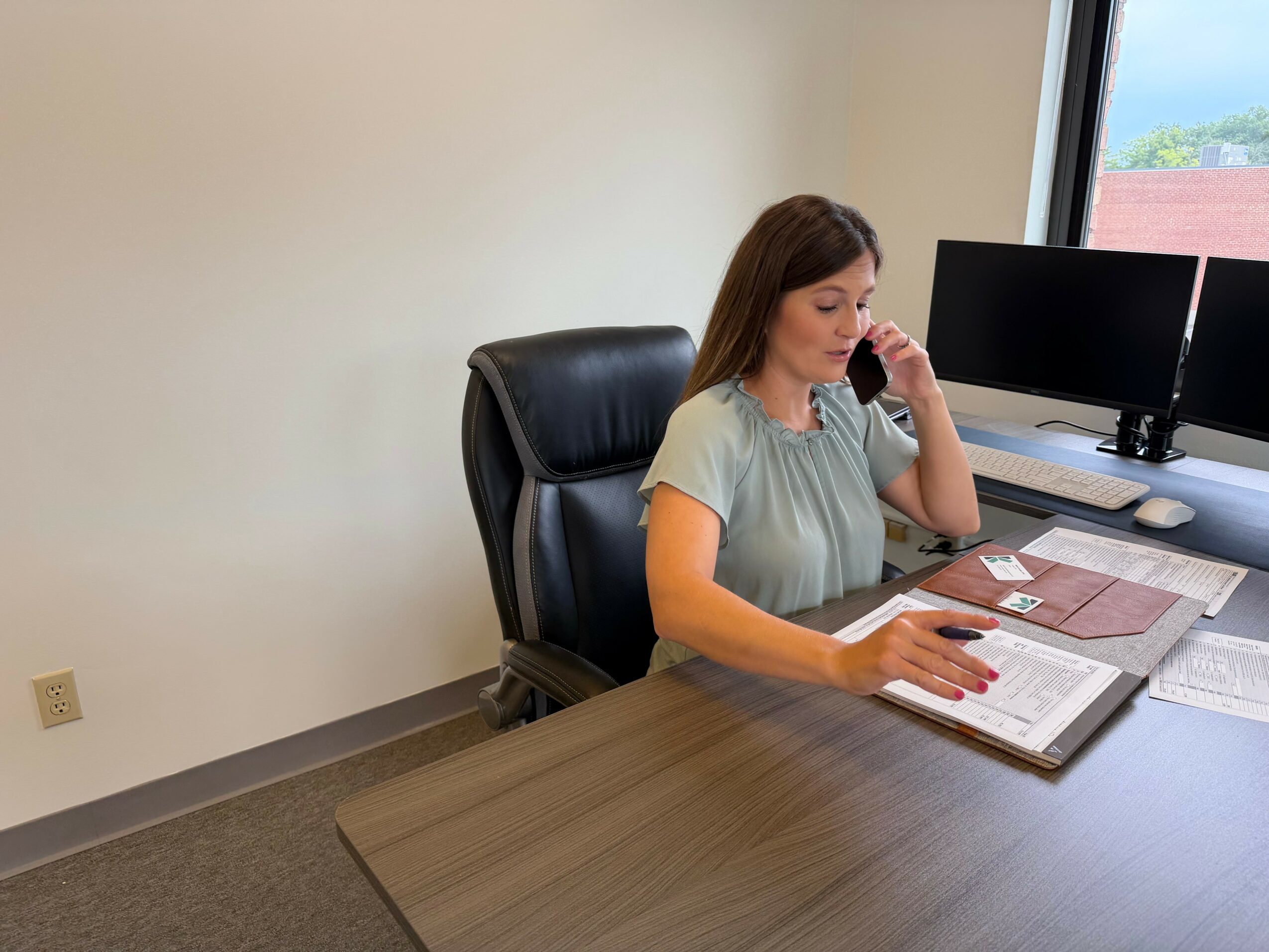 Woman sitting at desk, talking on phone, reading a book, with two monitors on desk, near window.