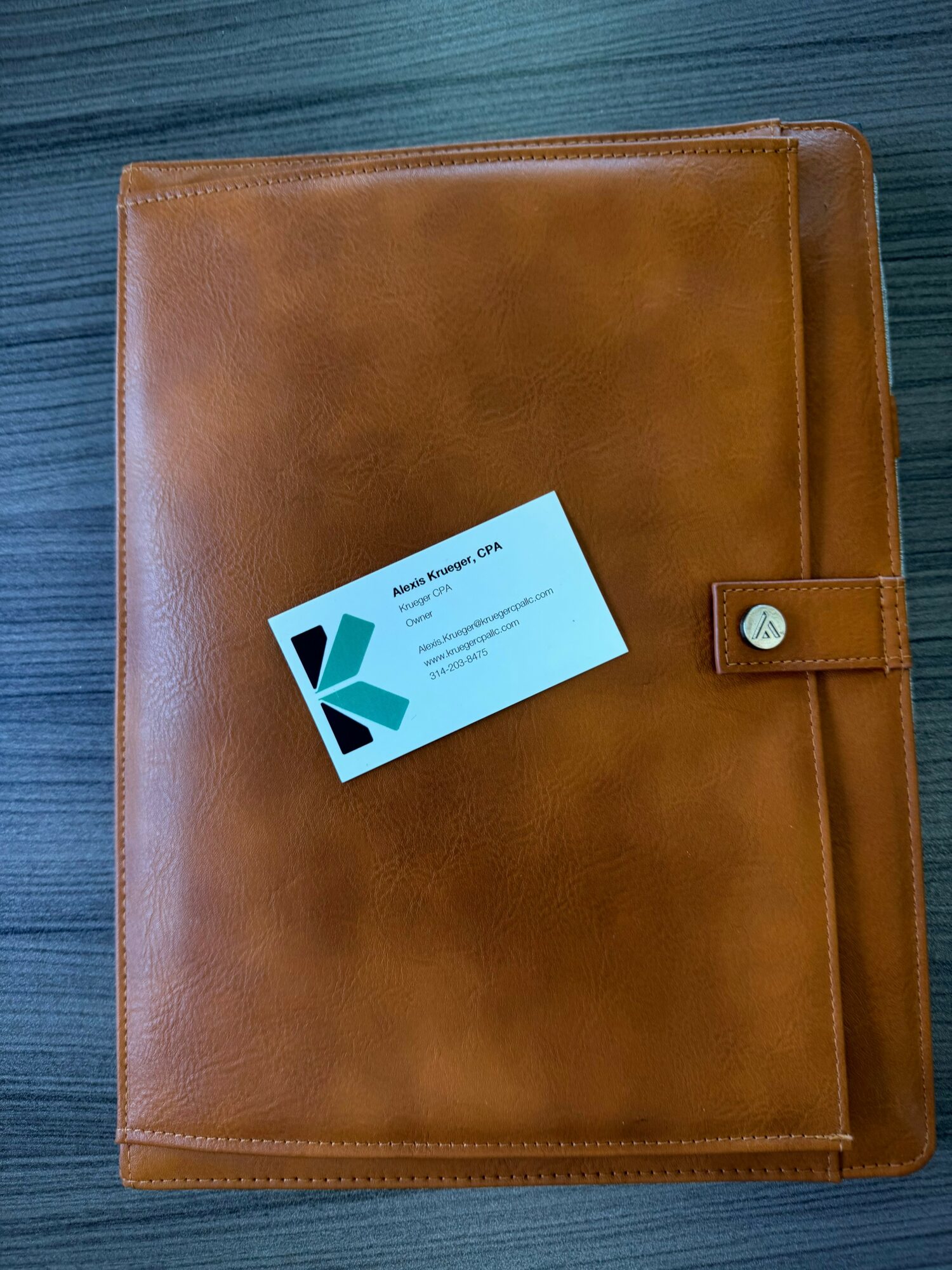 Brown leather-bound folder with a strap and button closure, placed on a wooden surface, with a business card on top.