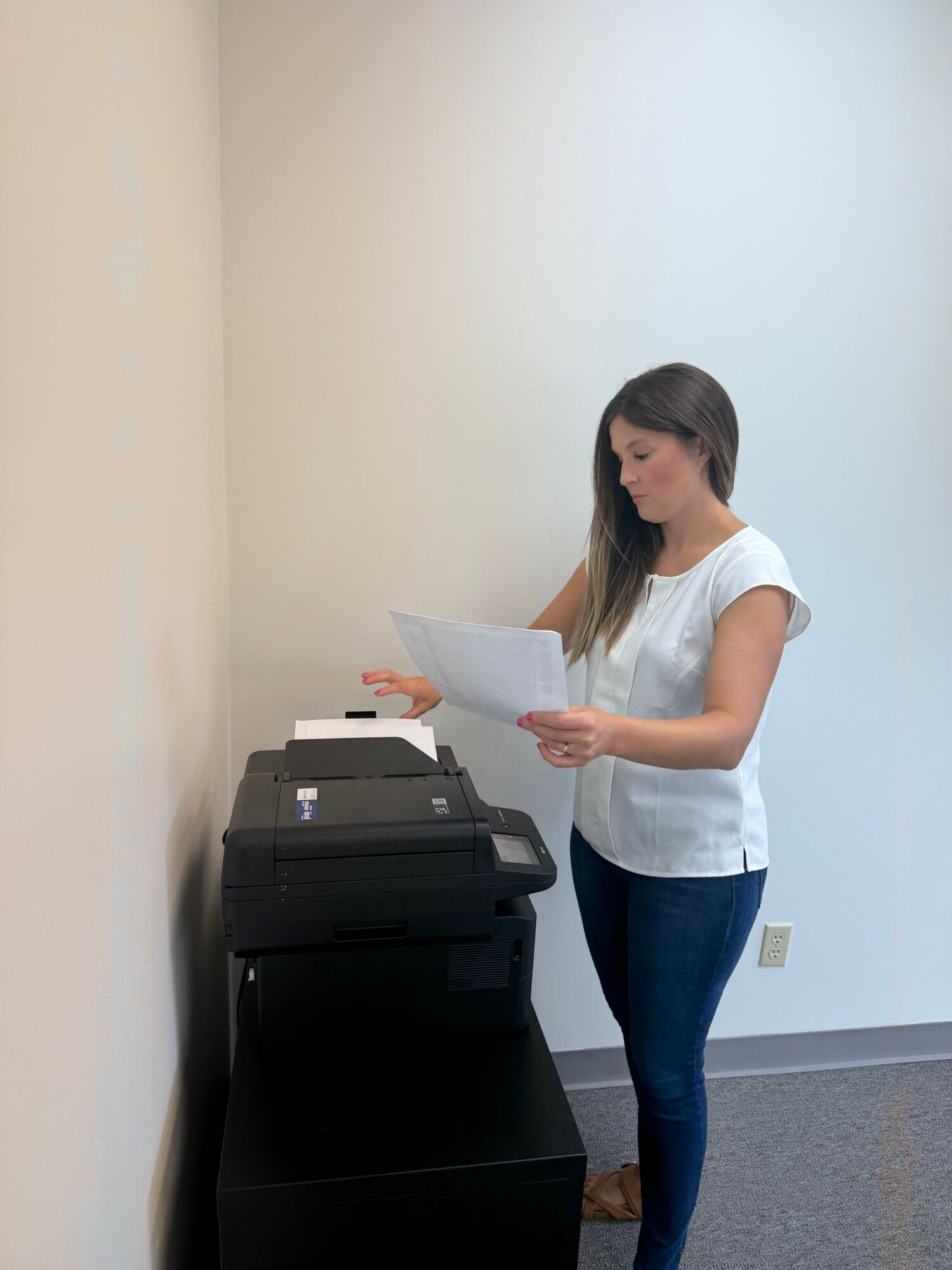 Woman using a printer in an office, standing and leaning slightly forward, with a plain wall background.