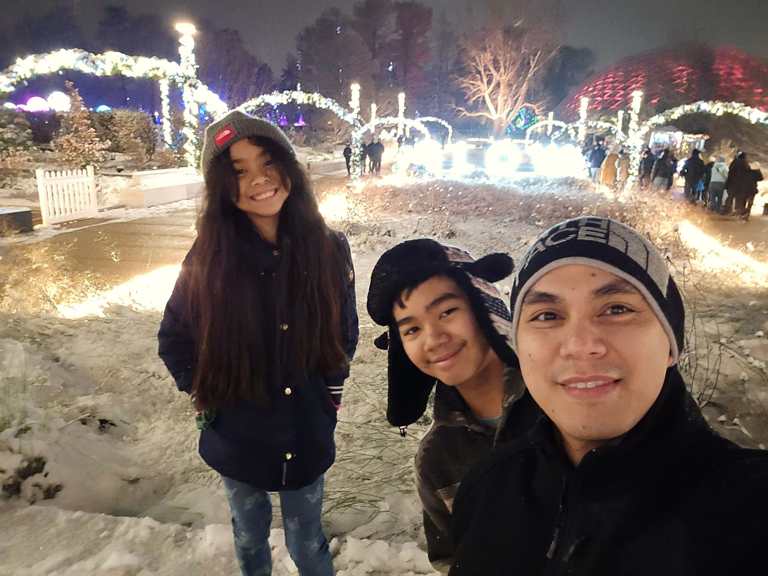 Three people standing outdoors at night with city lights and decorations in the background.