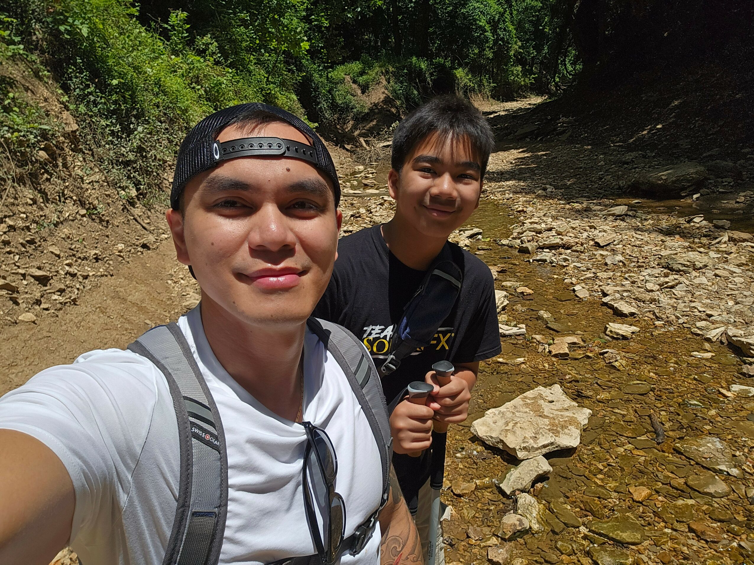 Two boys outdoors near a shallow stream with rocks, smiling at the camera, surrounded by green trees.