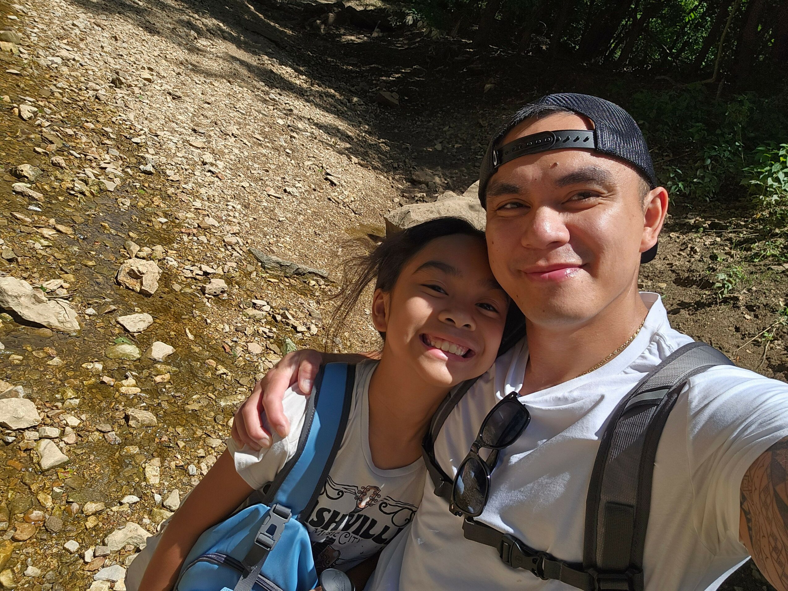 Two children outdoors, one wearing sunglasses and a cap, smiling, with a dirt path and greenery in the background.