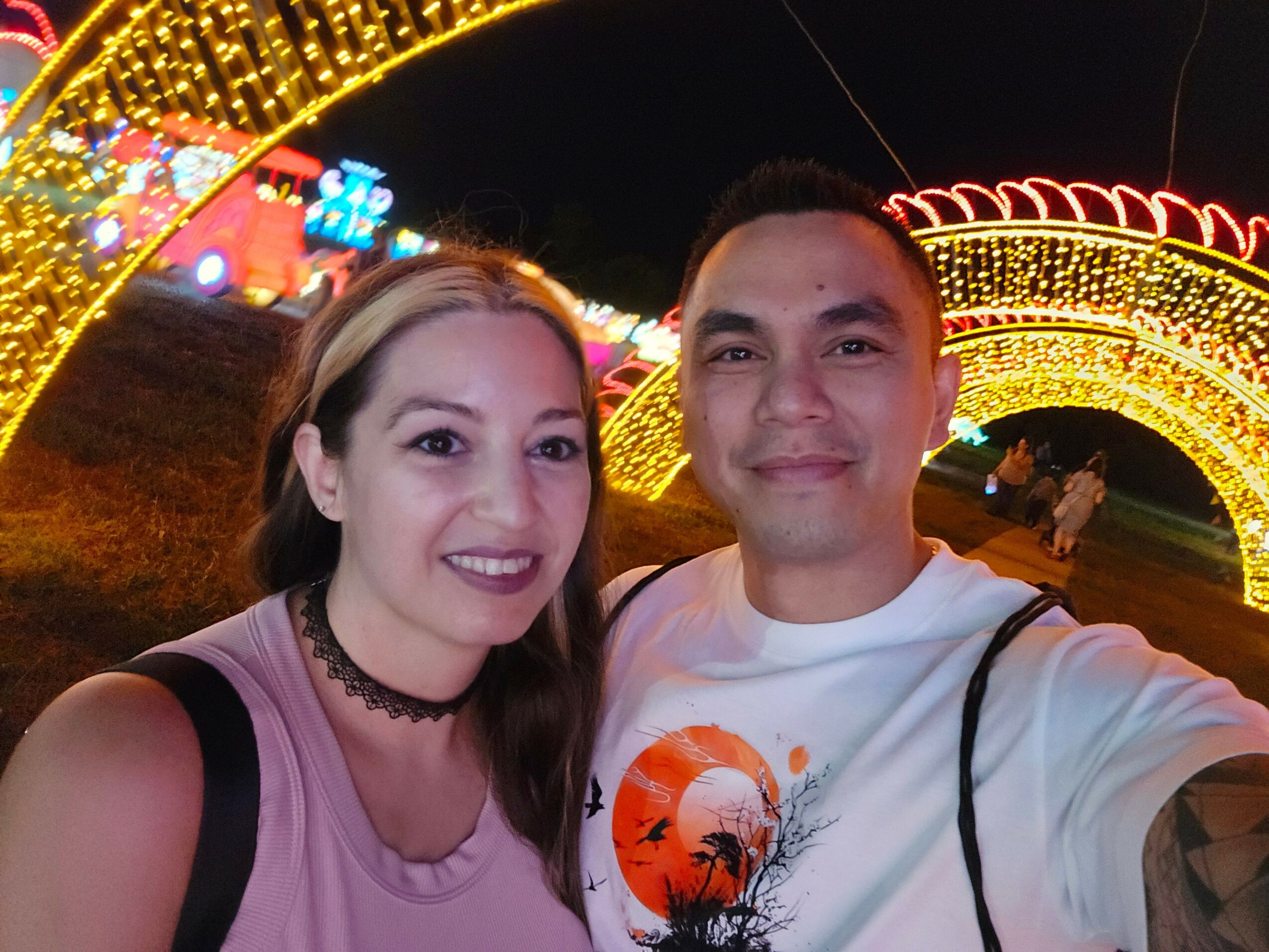 Two people smiling at night with colorful lights and decorations in the background.