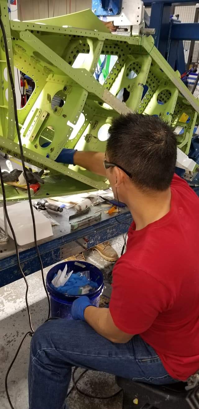Person working on a large green mechanical structure in a workshop, sitting on a stool with tools and a blue bucket nearby.