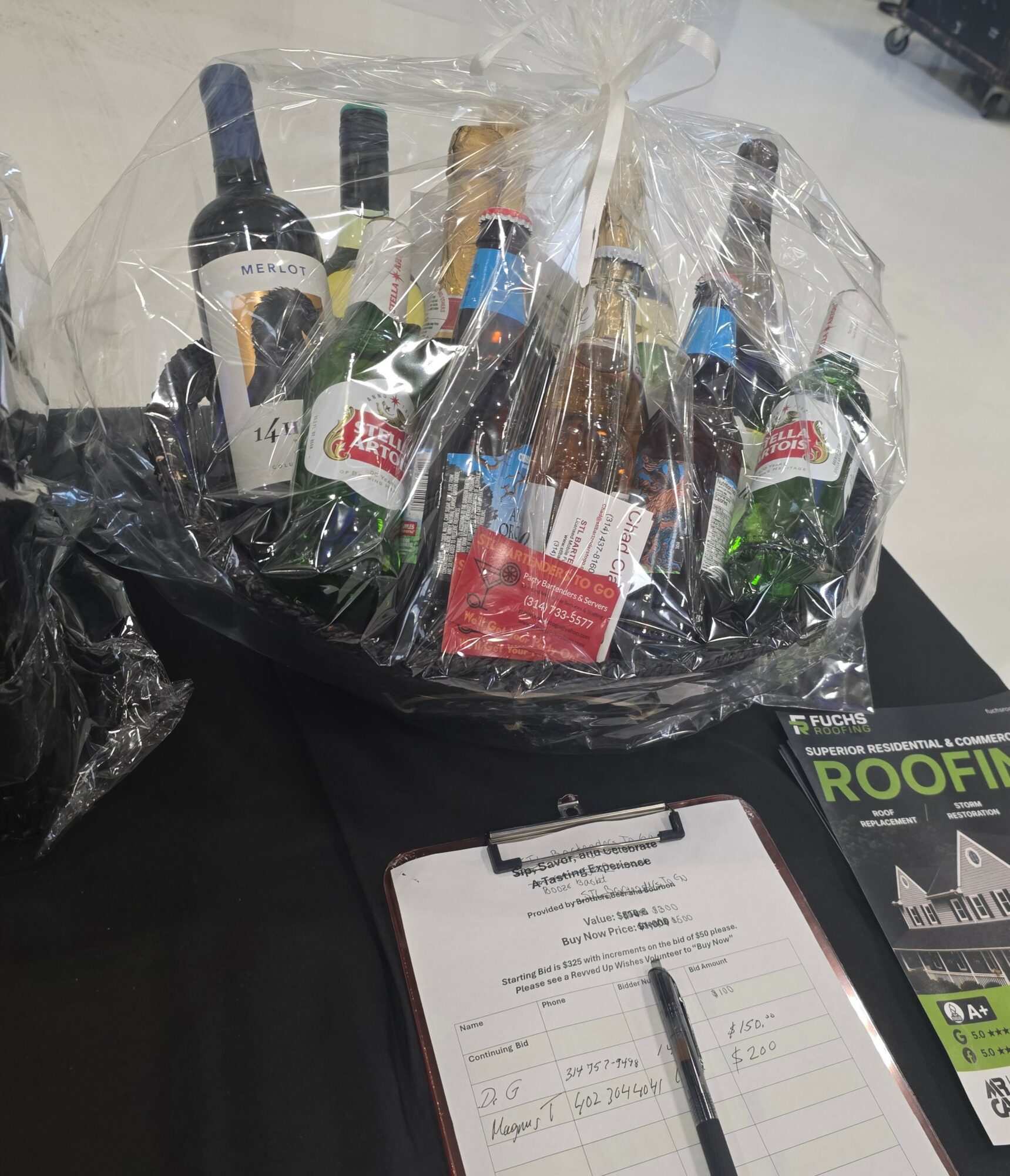 Gift basket with assorted bottles wrapped in clear plastic, placed on a black surface with a clipboard nearby.