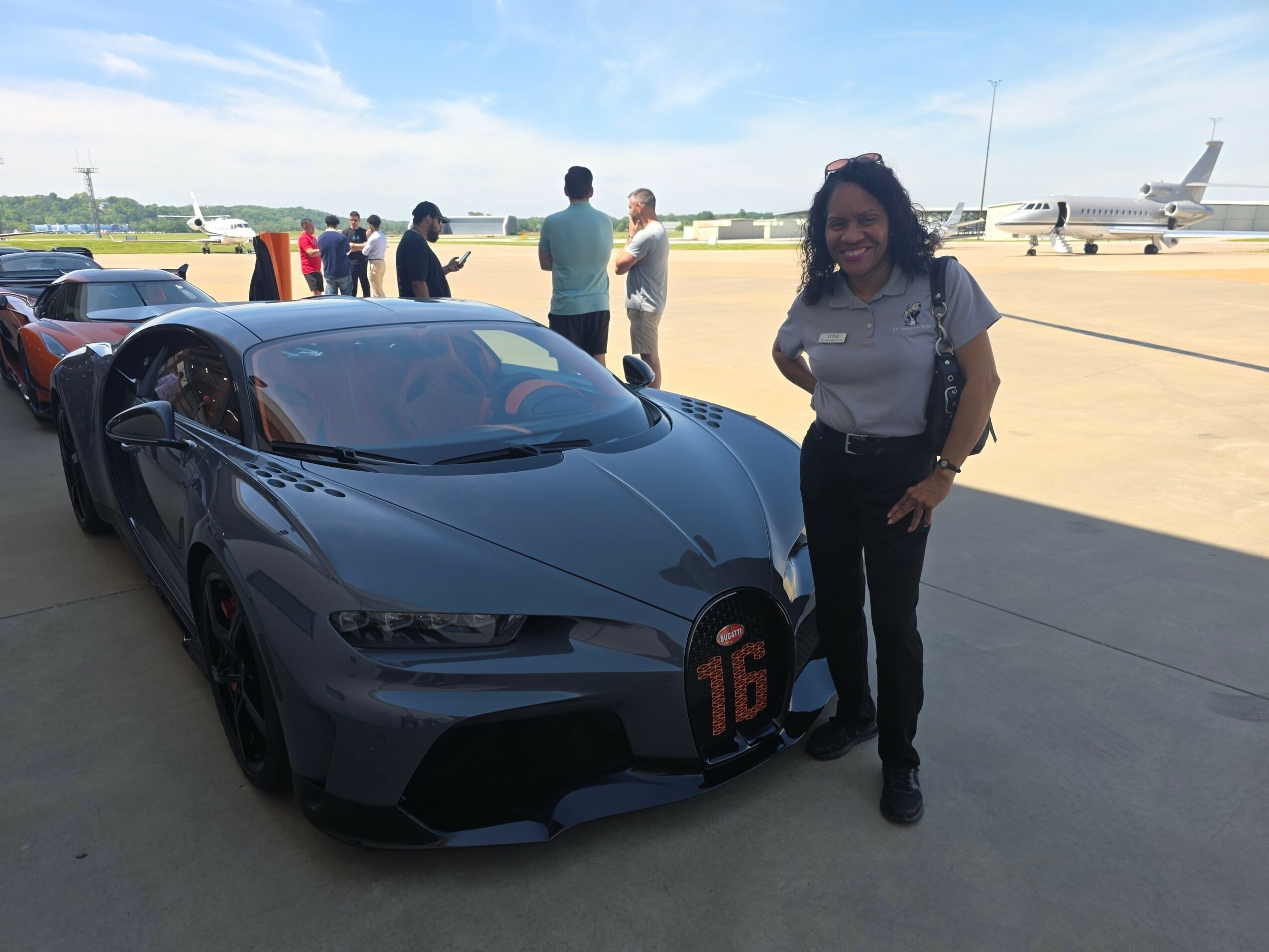 Woman standing next to a dark sports car at an airport tarmac with people and planes in background.
