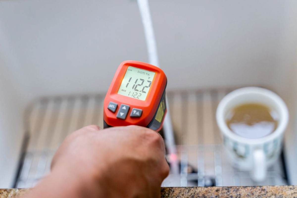 Hand holding a digital infrared thermometer displaying 122.4°F, with a cup of tea in the background on a table.