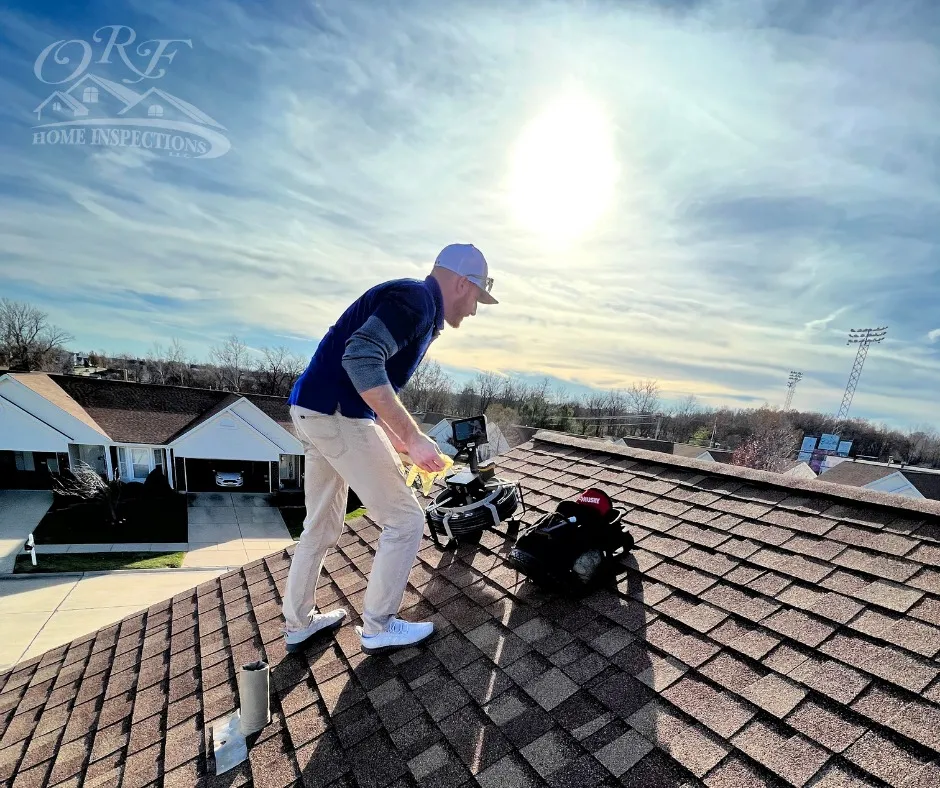 Person on roof with equipment, bright sun in sky, rooftops and trees in background, clear weather.