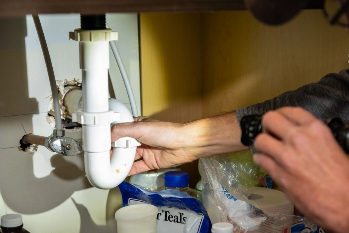 Person's hand reaching under sink pipe, with a hand holding a tool nearby, in a kitchen or bathroom setting.