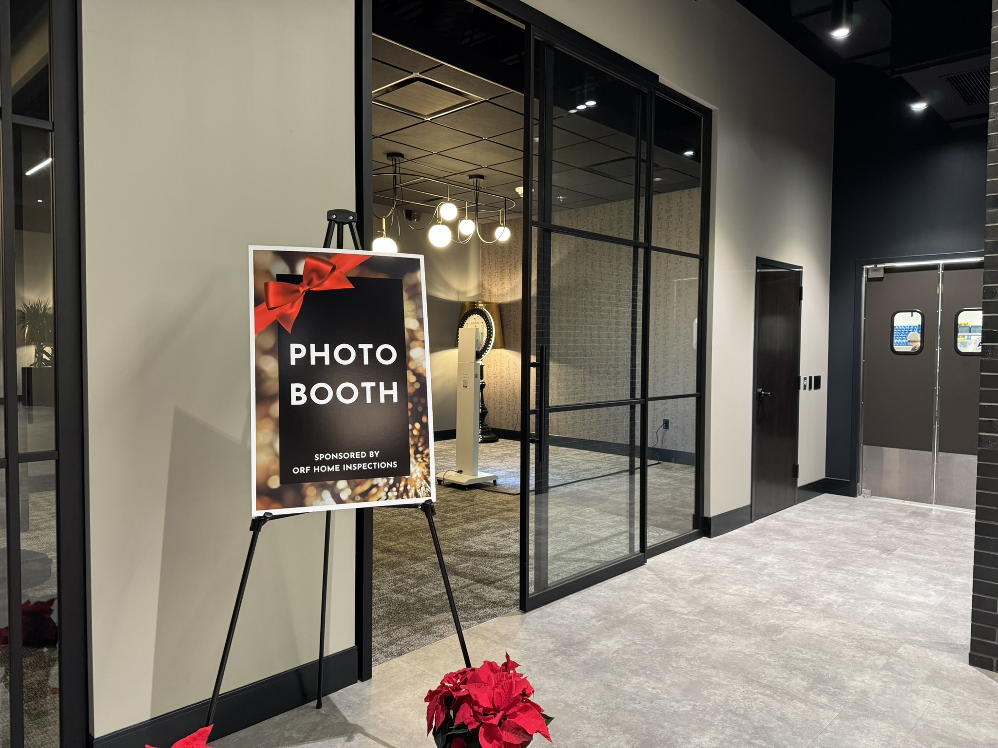 Photo booth sign with red bow and poinsettia flowers in a modern indoor space, glass walls, and elevator doors in background.