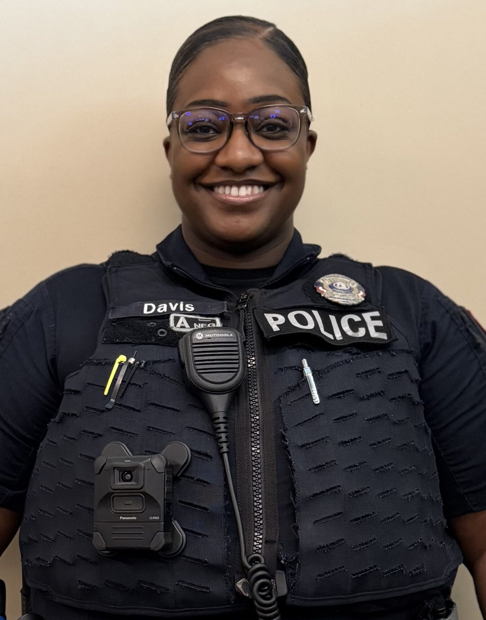 Smiling police officer wearing glasses and uniform with badge and radio, standing against a plain background.