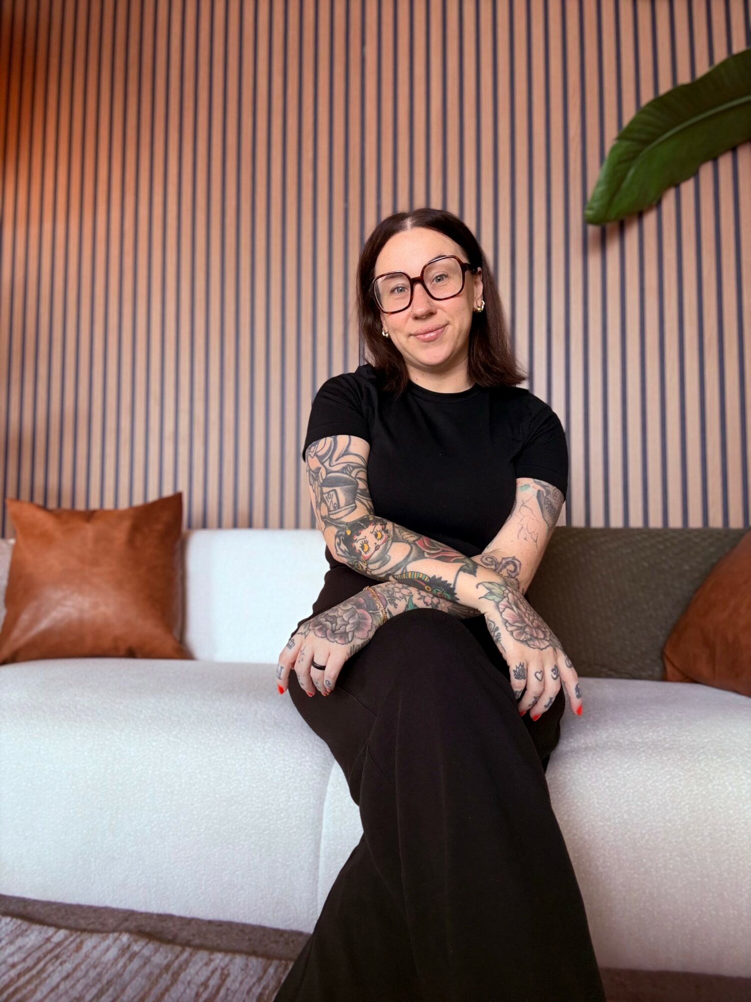 Woman with glasses and tattoos sitting on a sofa in front of a striped wall, with large green leaf nearby.