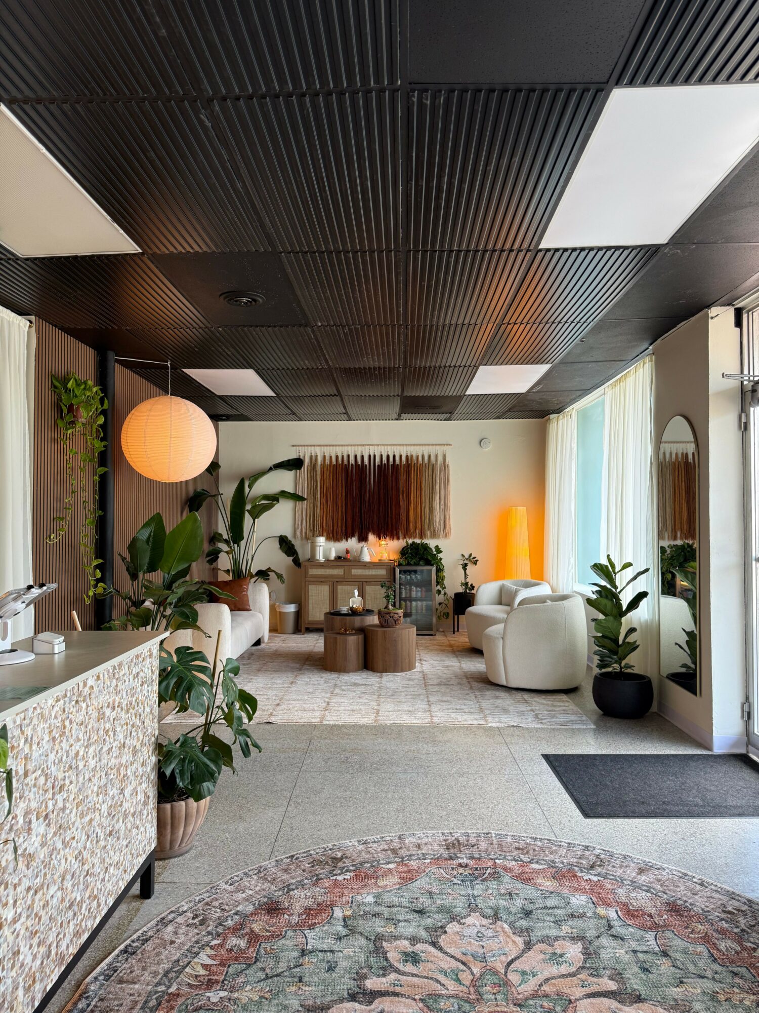 Living room with white chairs, potted plants, a round rug, and a large window with curtains.