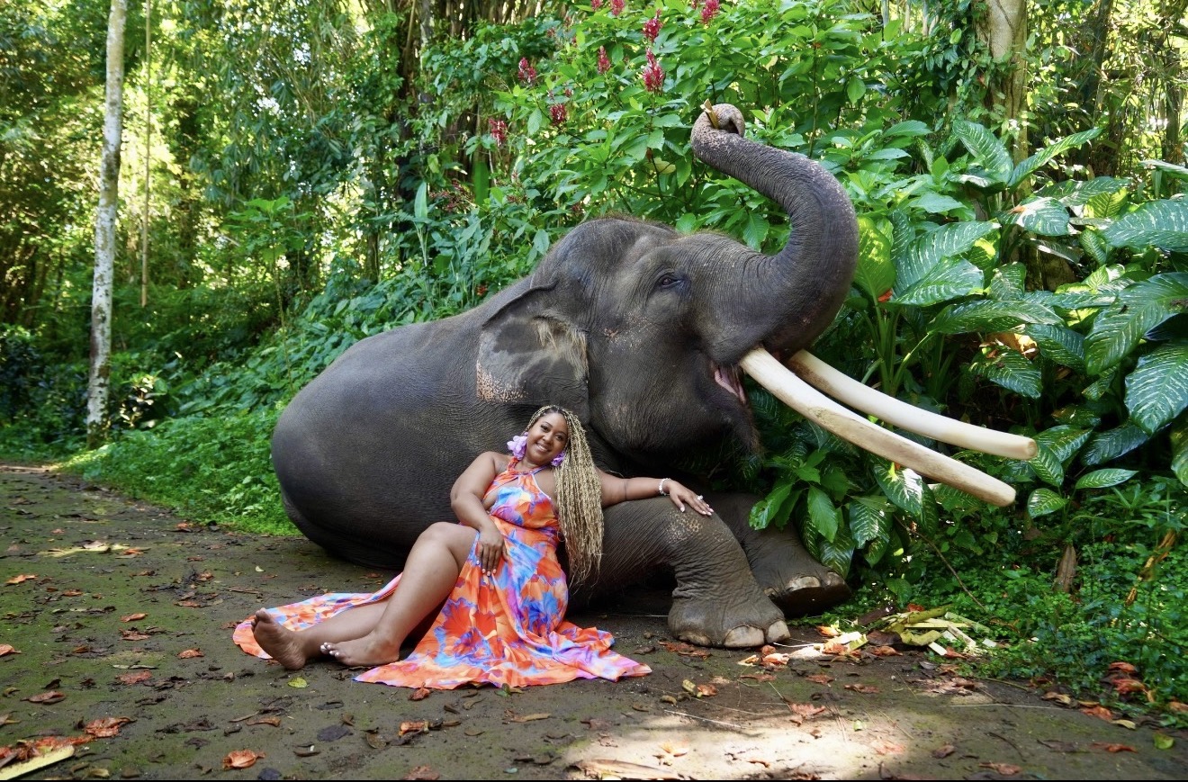 Woman in colorful dress sitting on ground next to a large elephant lying in a lush green forest.