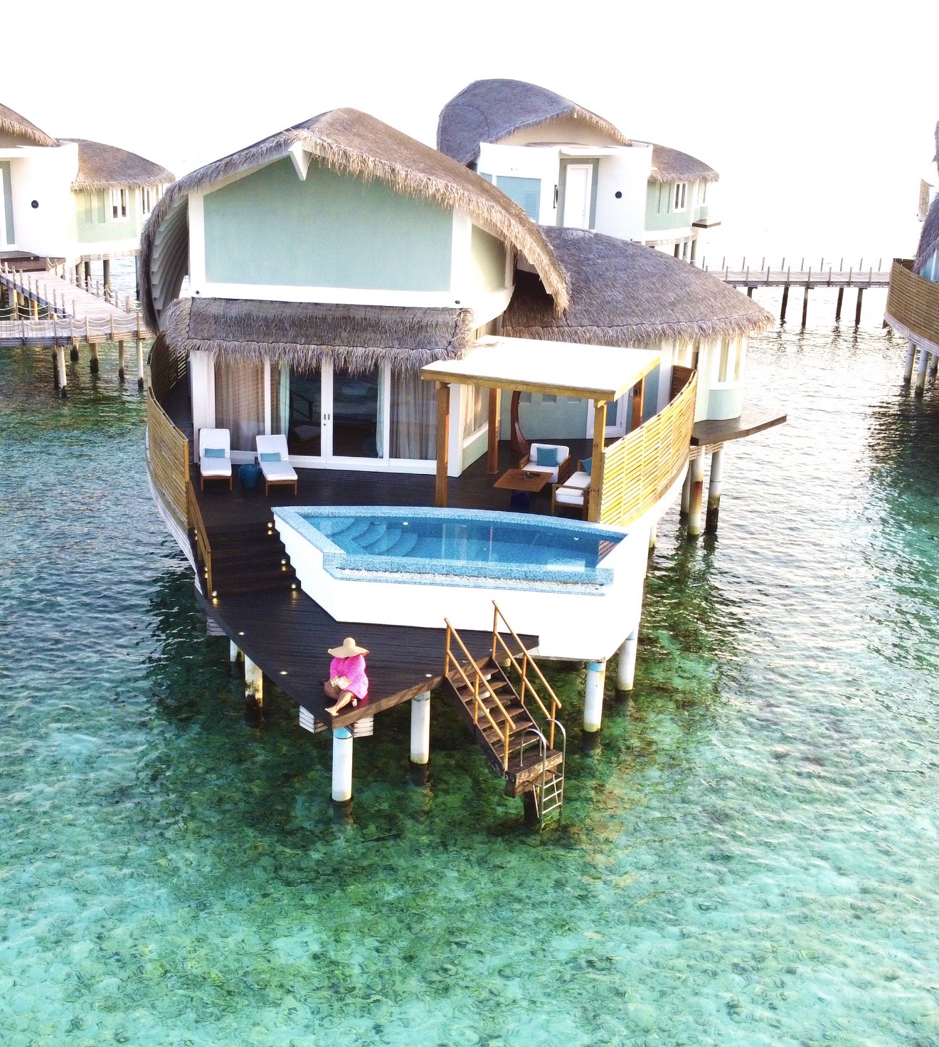 Overwater bungalow with a swimming pool, deck, and stairs leading to water, set over clear turquoise water.