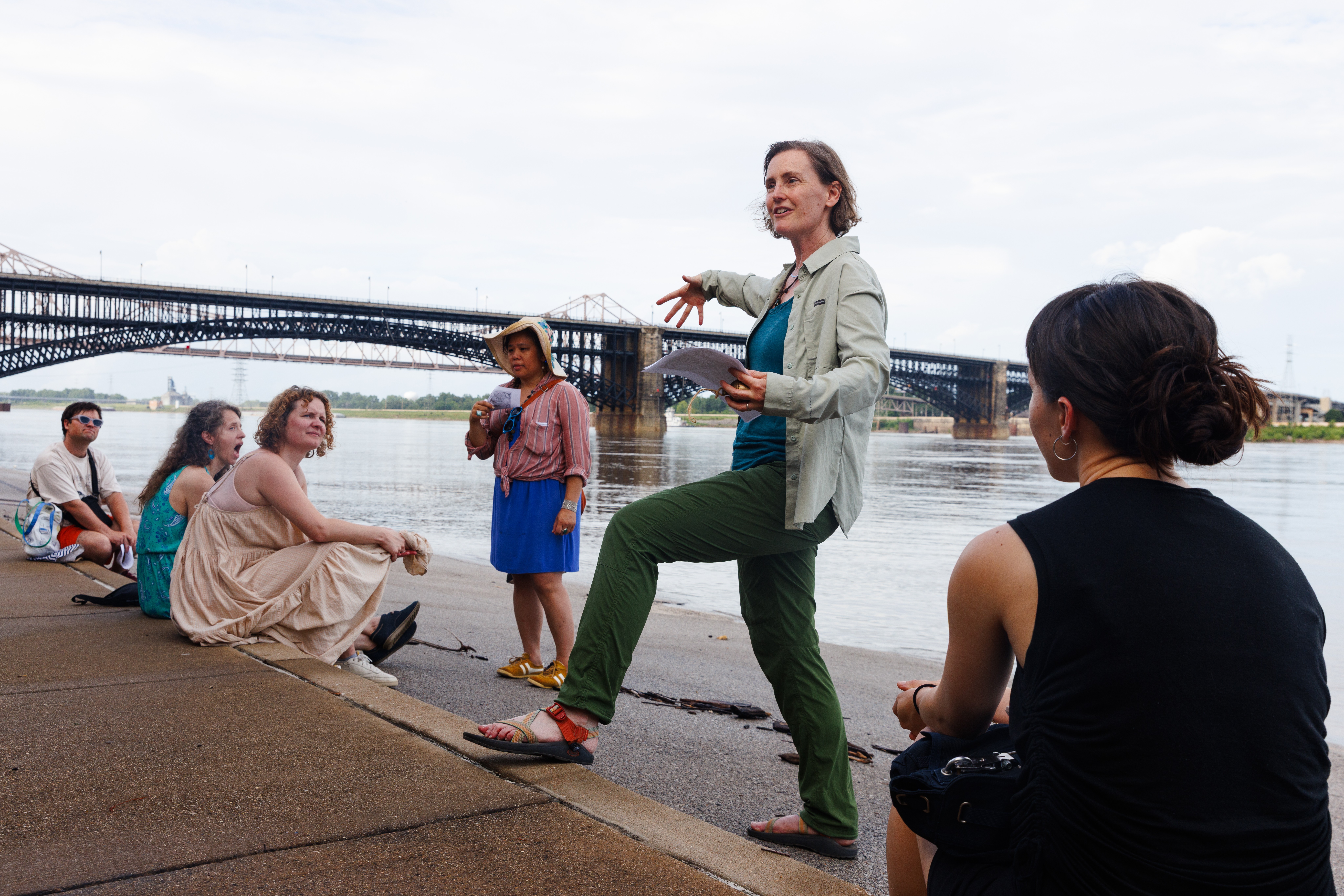 Group of people by a riverbank with a bridge in the background, woman speaking to the group, some sitting and standing.