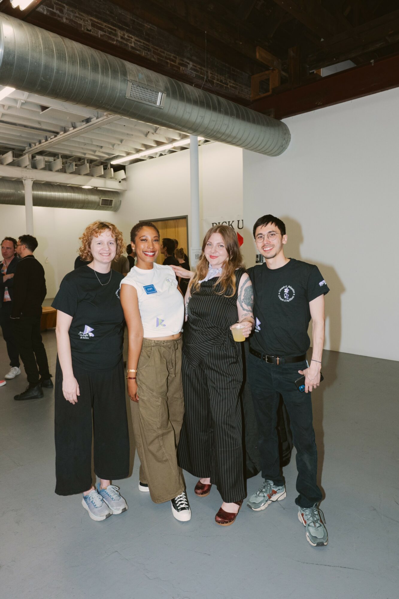 Four people standing together indoors, smiling, with industrial ceiling and ductwork visible above.