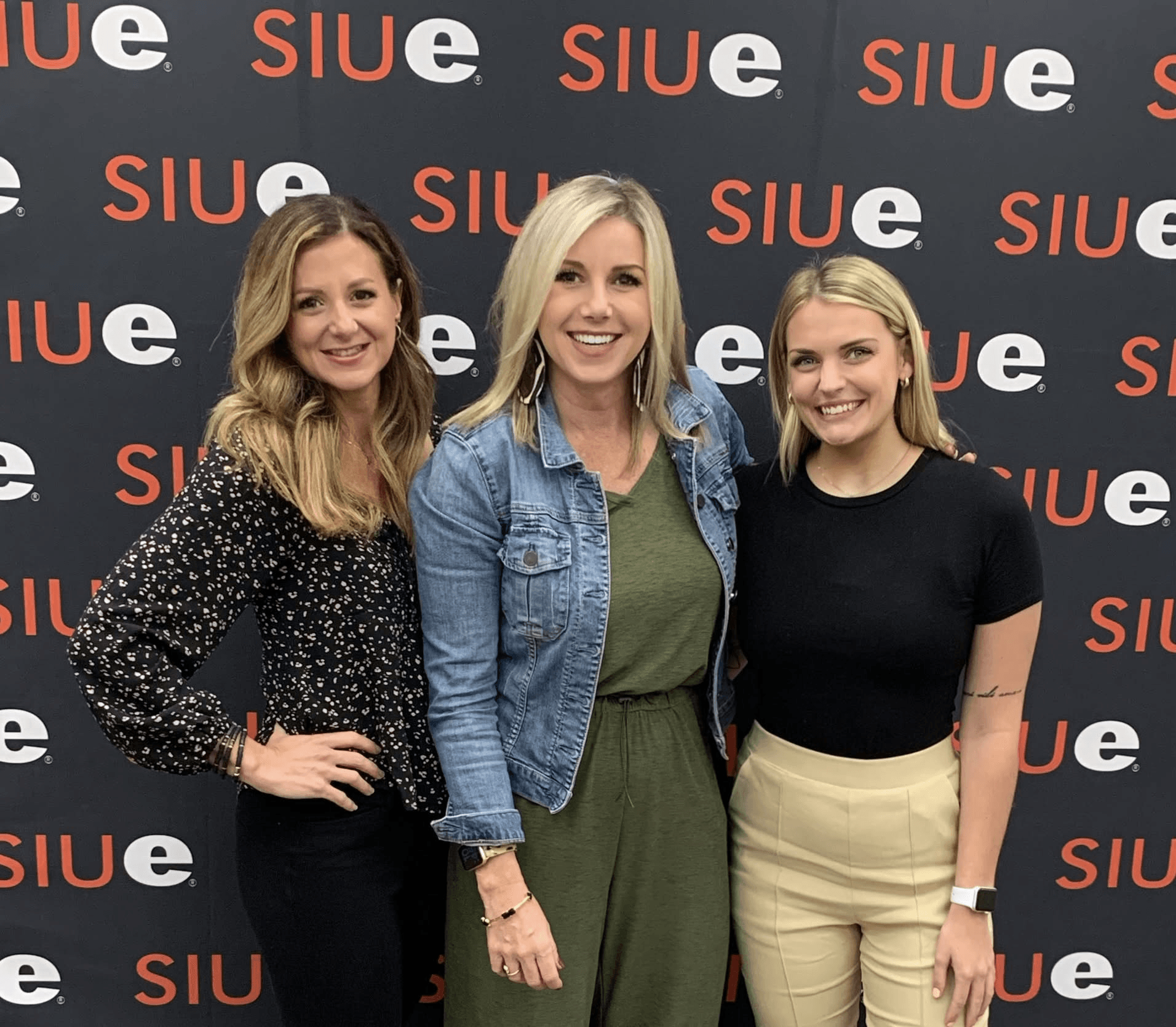 Three women standing together in front of a black backdrop with red and white 'SIUE' logos, smiling at the camera.