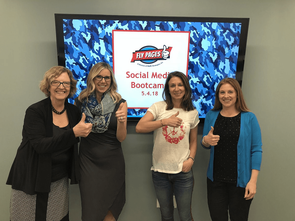 Four women standing in front of a screen displaying a logo and text, smiling and giving thumbs up.
