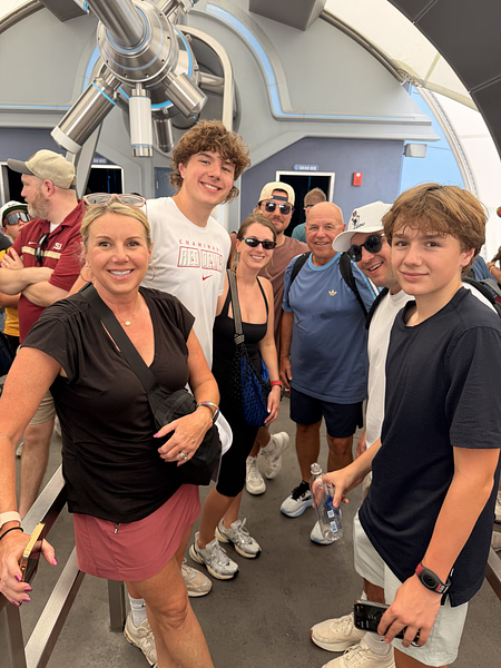 Group of people smiling inside a science or space-themed exhibit with a large telescope-like structure in the background.