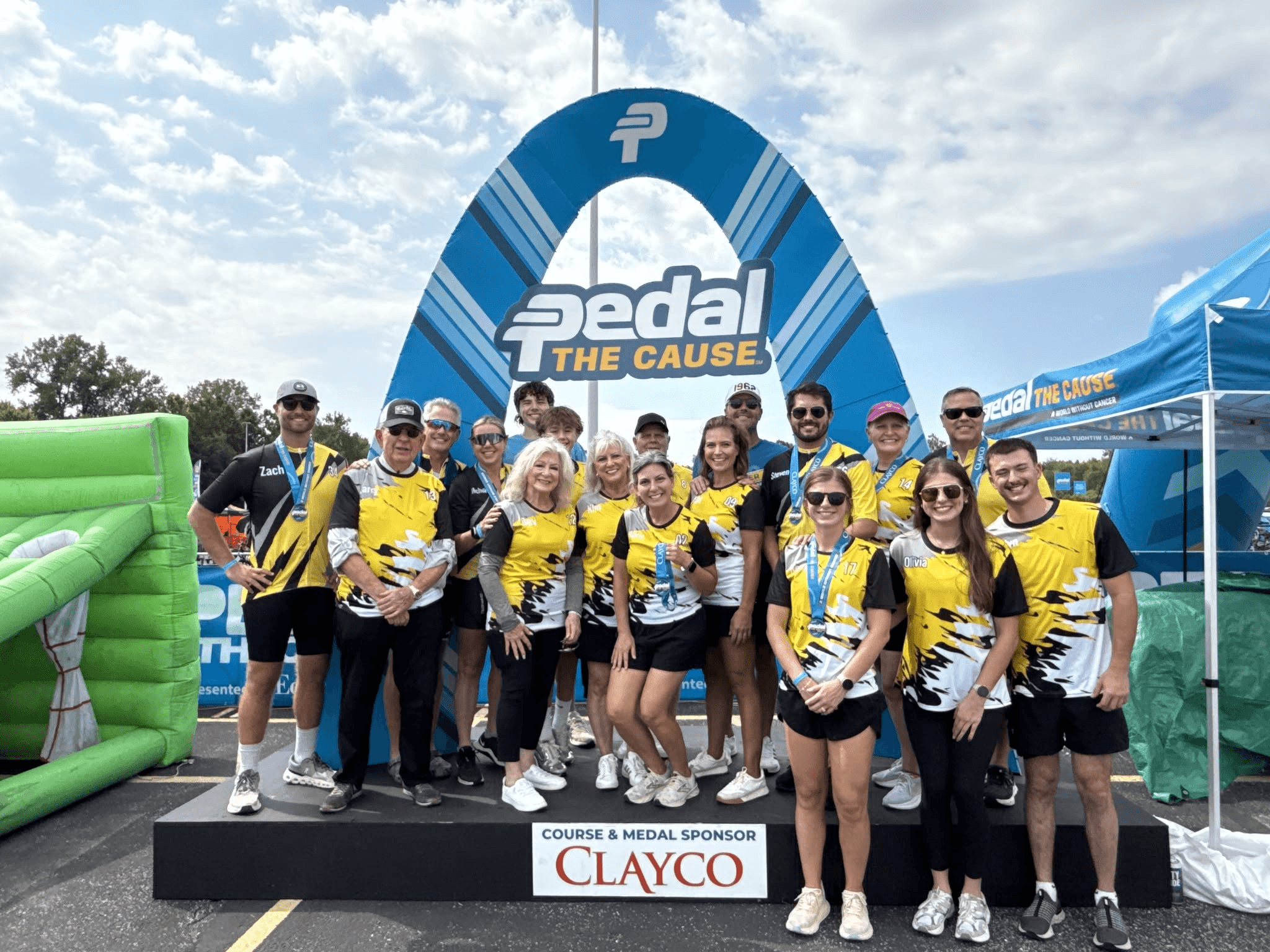 Group of people in yellow and black shirts standing on a podium with a large blue arch and sky background.