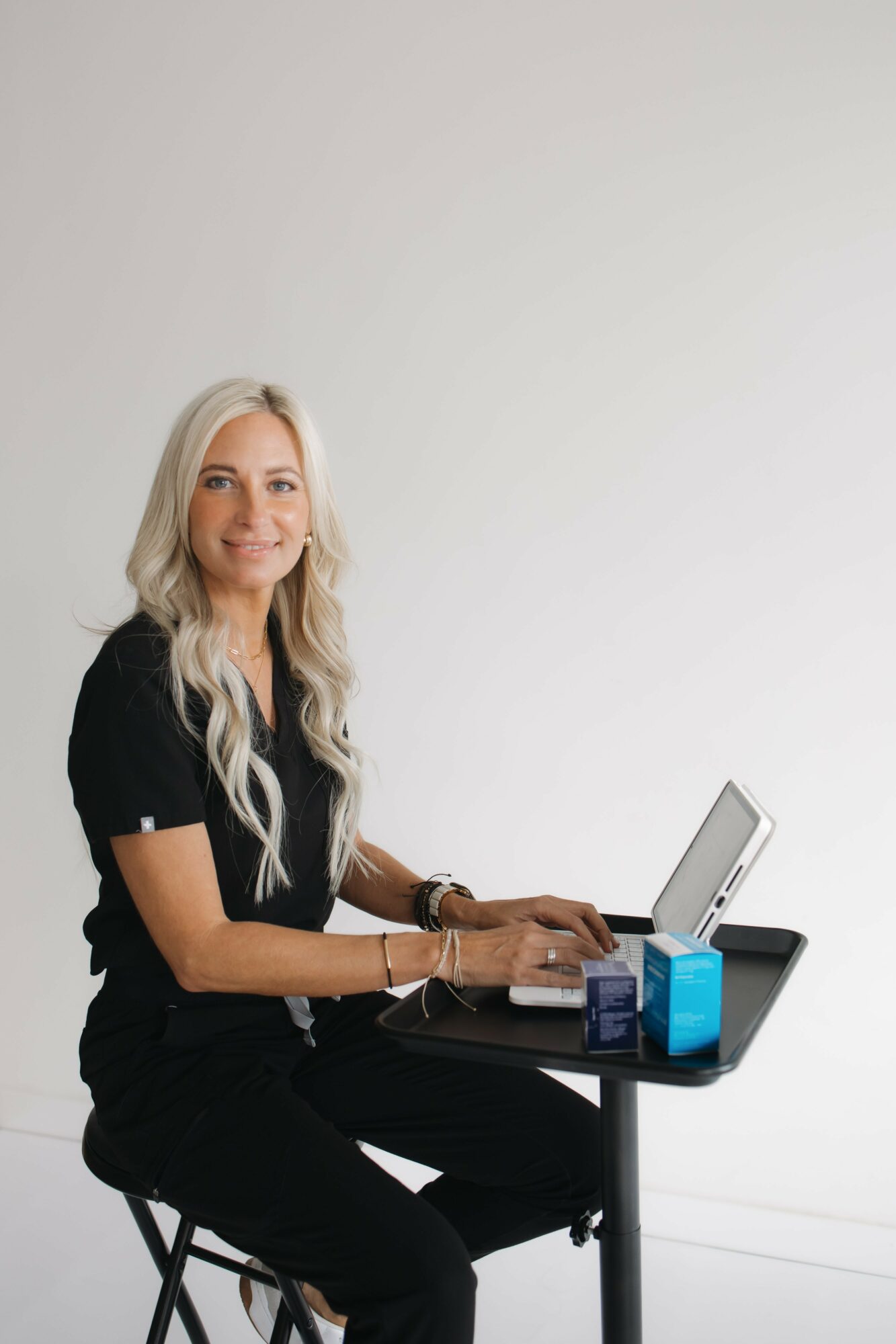 Woman with long blonde hair sitting at a black table using a laptop, with a blue container nearby.