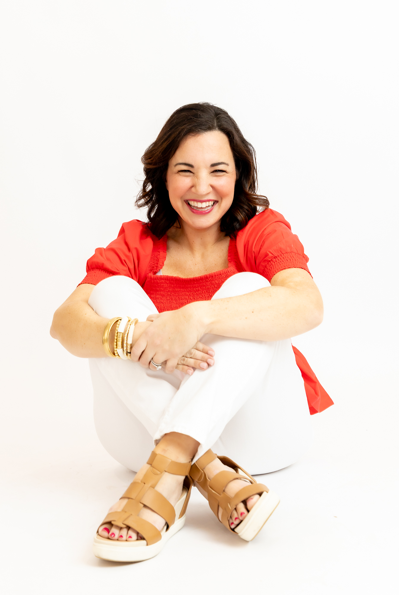 Smiling woman sitting with arms around knees, wearing a red top, white pants, and tan sandals, against a white background.