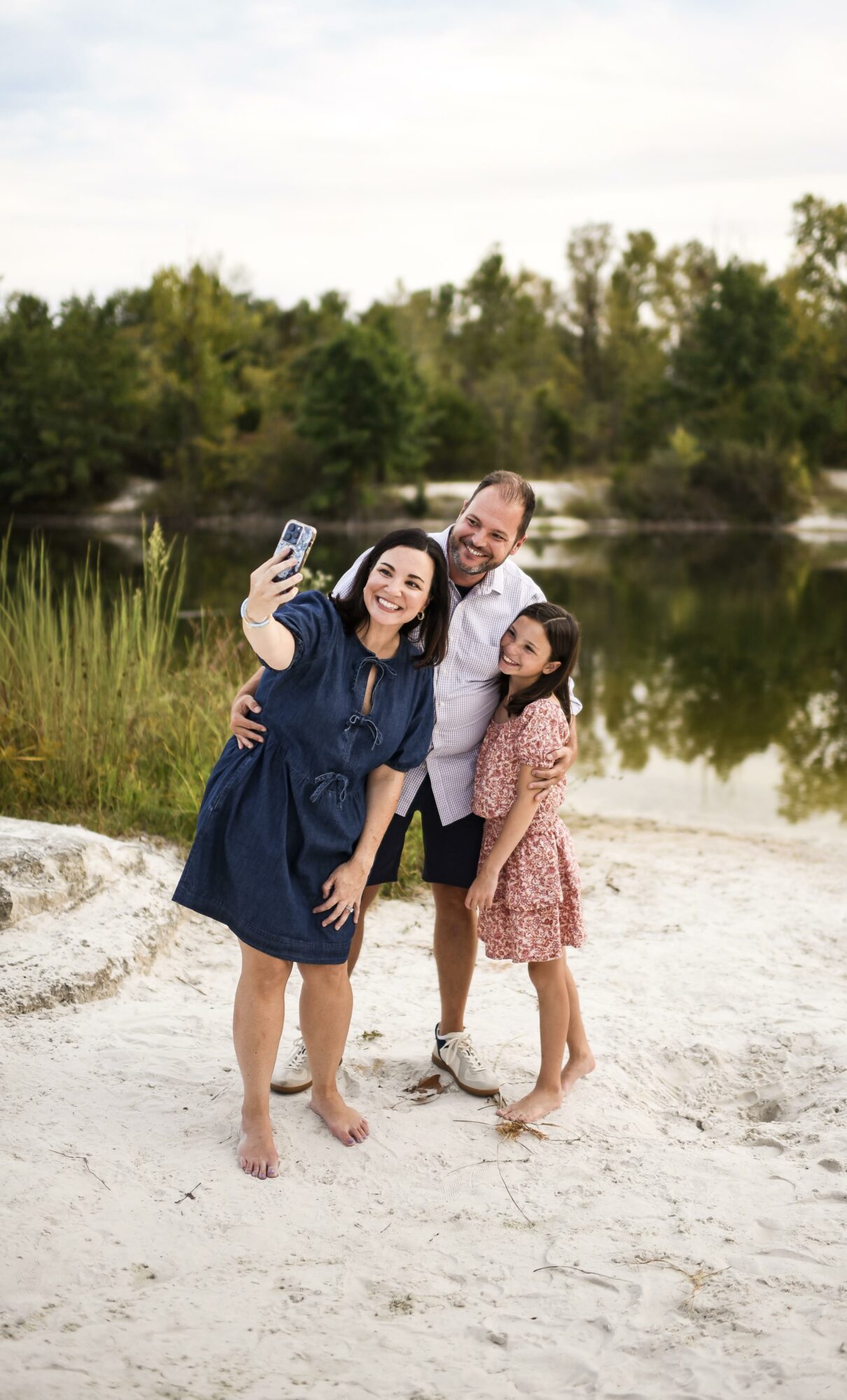Family of three taking a selfie on a sandy beach near a river, with trees in the background.