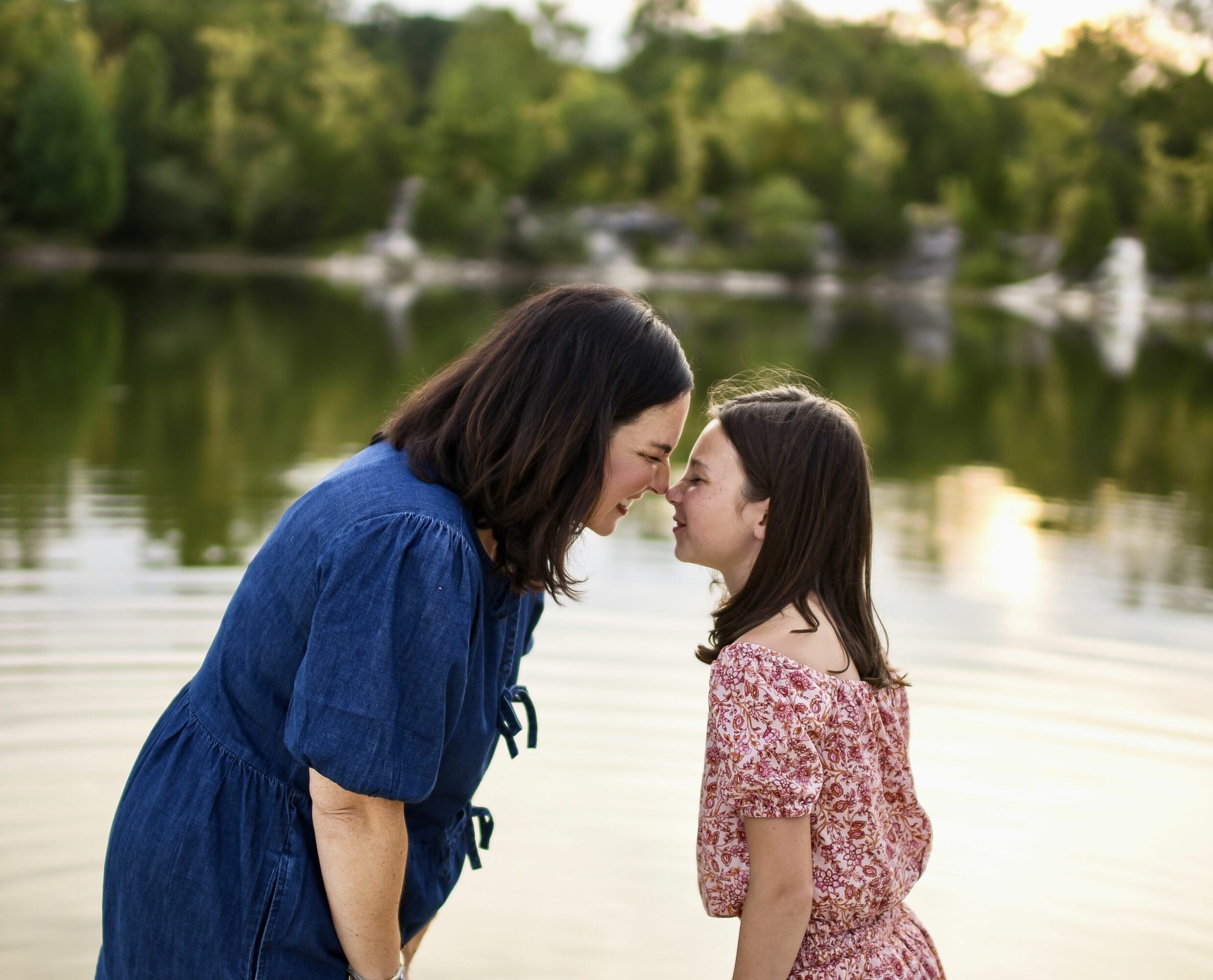Woman and girl face each other near a lake, smiling, with trees in the background.