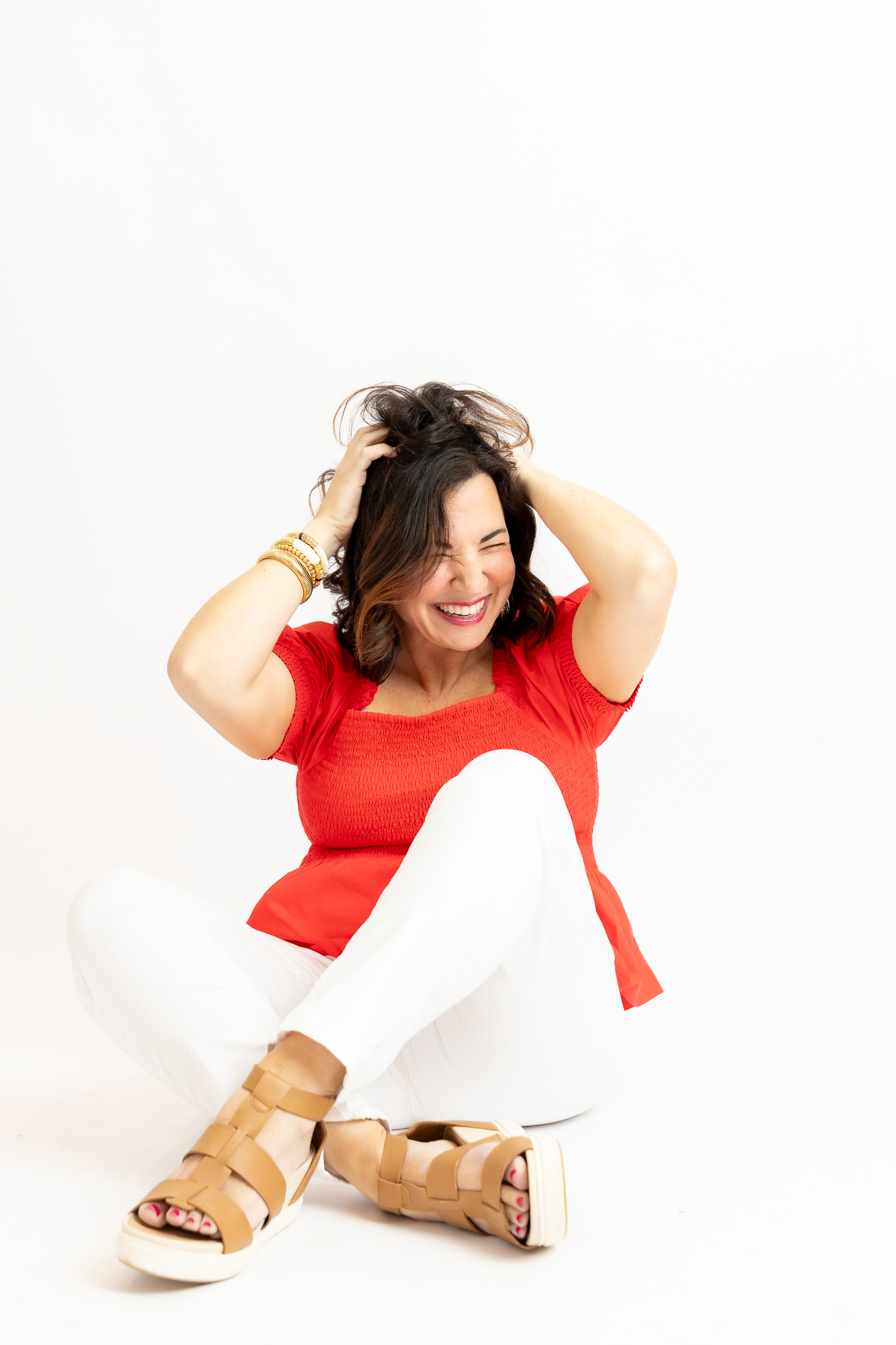 Woman sitting on the floor, smiling, wearing a red top, white pants, and tan sandals, with hands in her hair.
