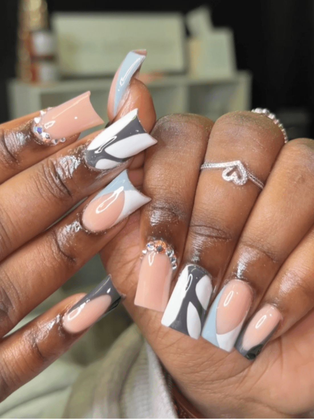 Close-up of hands with manicured nails in pastel pink, white, gray, and black with abstract designs, and a silver ring with a heart.