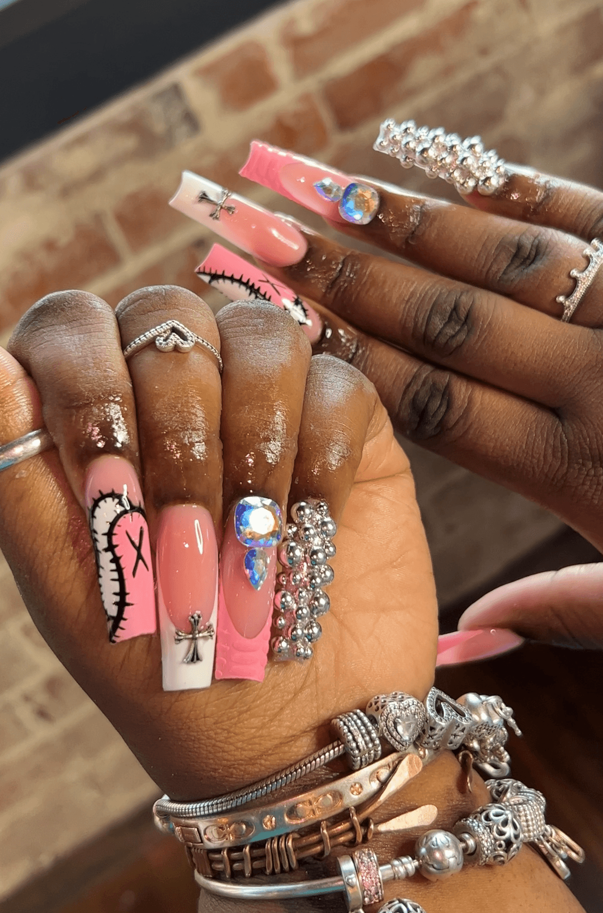 Close-up of hands with long, decorated nails and multiple rings, set against a brick wall background.