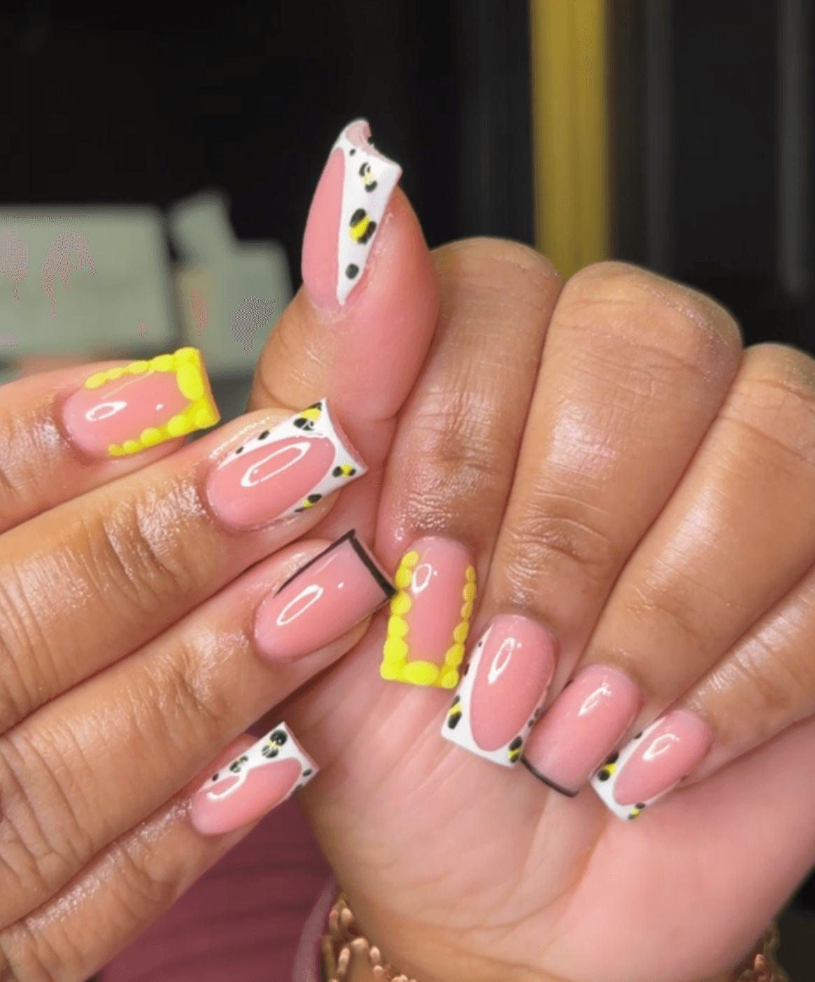 Close-up of hands with colorful, decorated nails featuring pink, yellow, white, and black designs, some with polka dots and yellow borders.