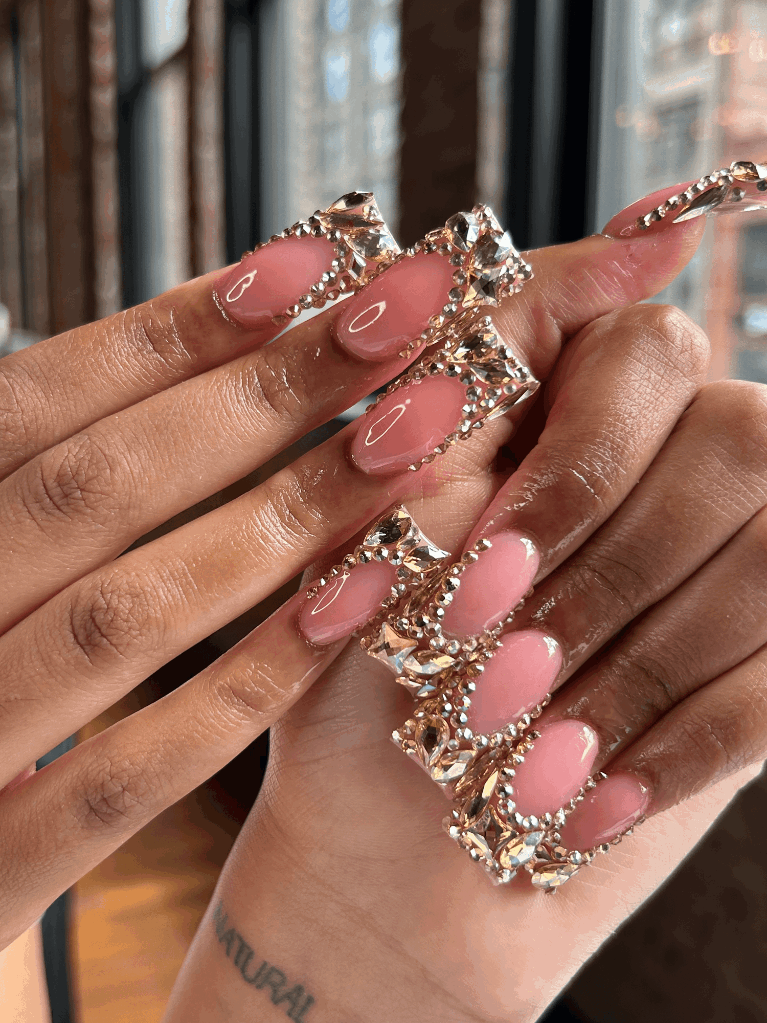Hands with long, decorated nails and gold jewelry, in front of a window with a cityscape background.